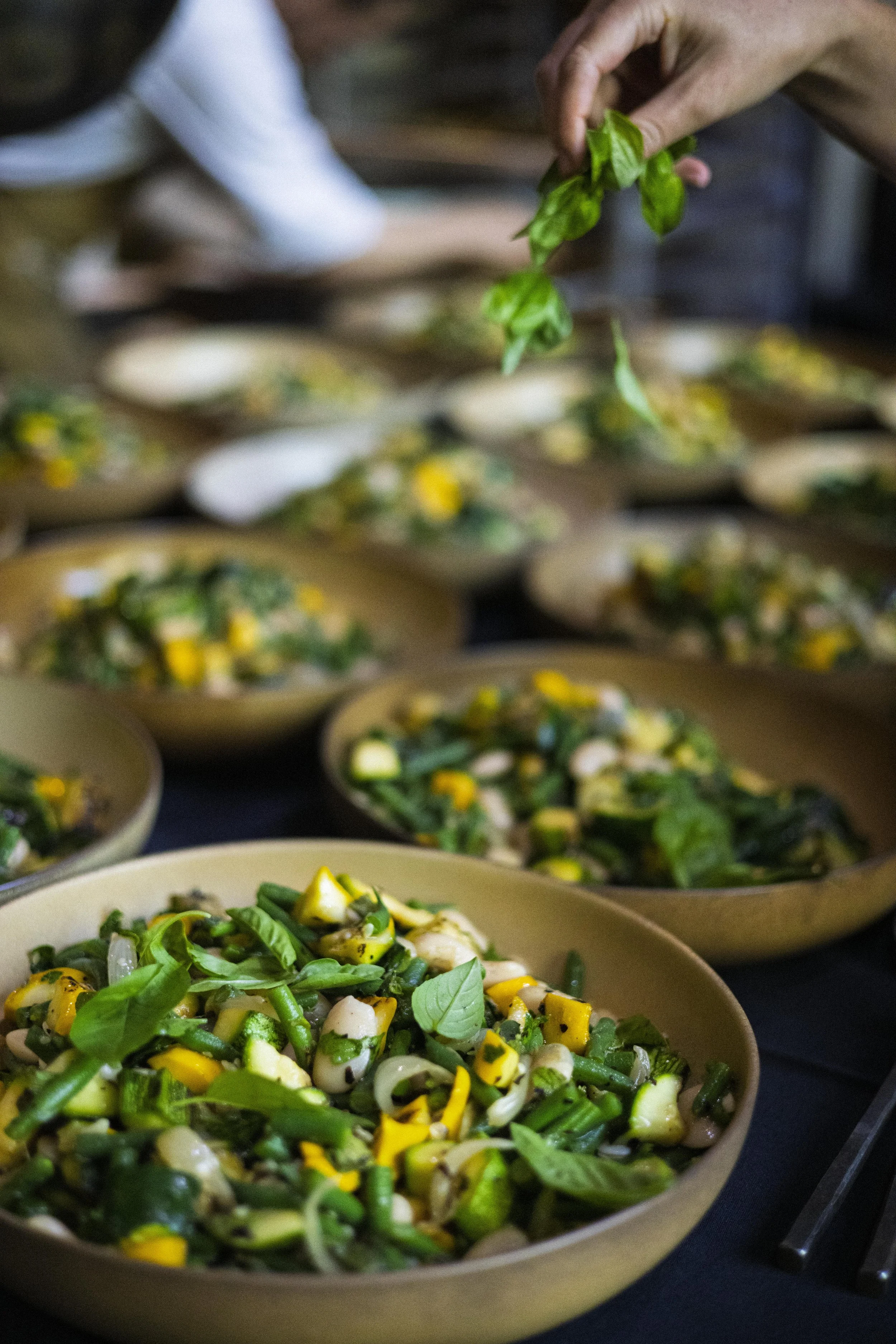 A person adding fresh basil leaves to a bowl of mixed vegetables, with multiple bowls of salad on a table, at a food service or catering event.