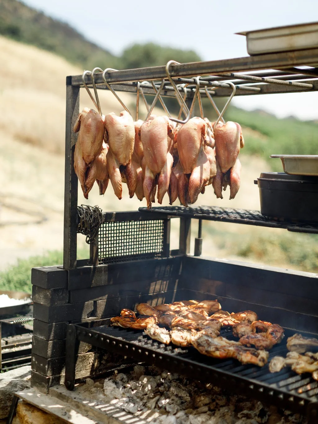 A barbecue grill outdoors with raw chickens hanging on hooks and cooked chicken pieces on the grill.