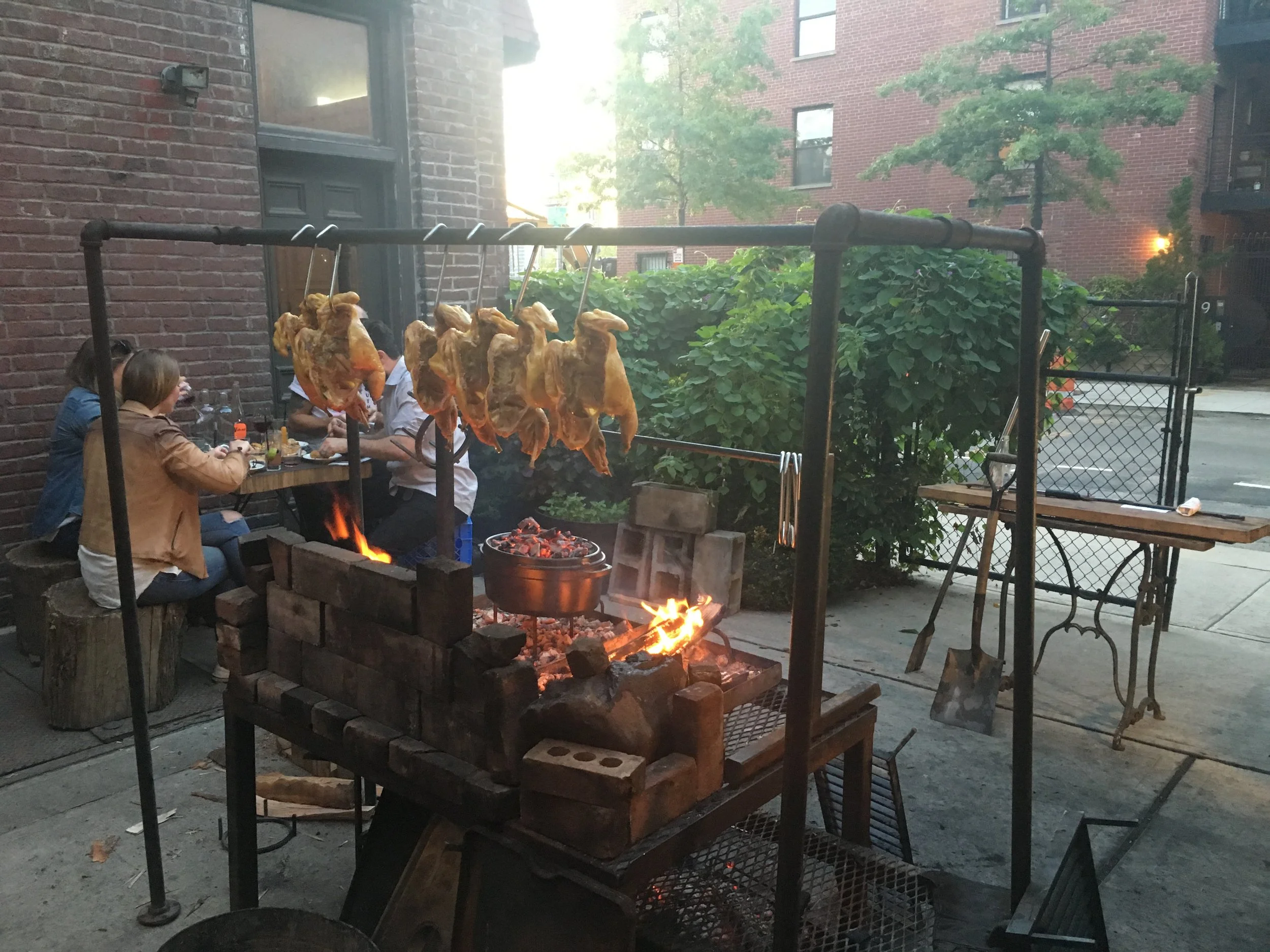 Outdoor scene of a backyard with a brick building in the background. There is a fire pit with burning wood and a grill with hot coals. Several chickens are hanging on hooks above the fire. A group of people are sitting at a table, eating and talking.