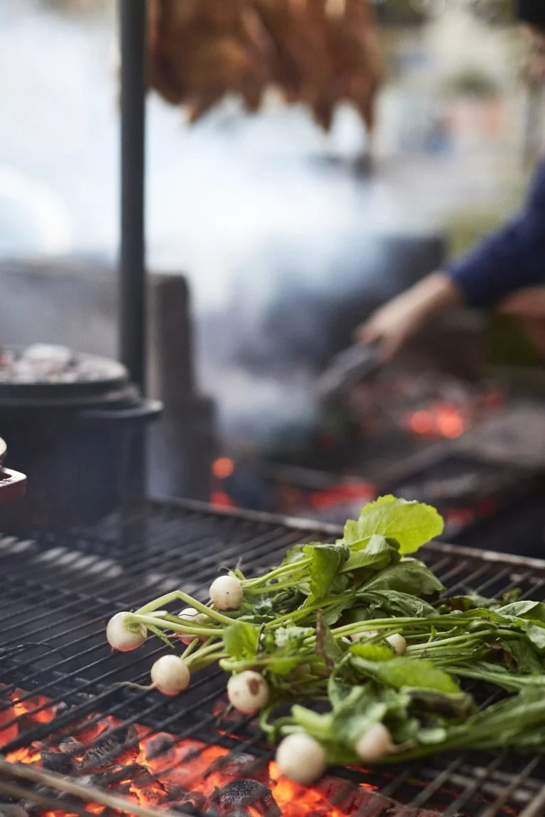 Green leafy vegetables, possibly radishes, grilling on an outdoor barbecue with flames beneath the grill.