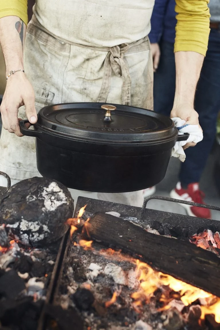 Person holding a black cast iron slow cooker over a campfire with glowing logs and flames, wearing an apron and yellow sleeves.
