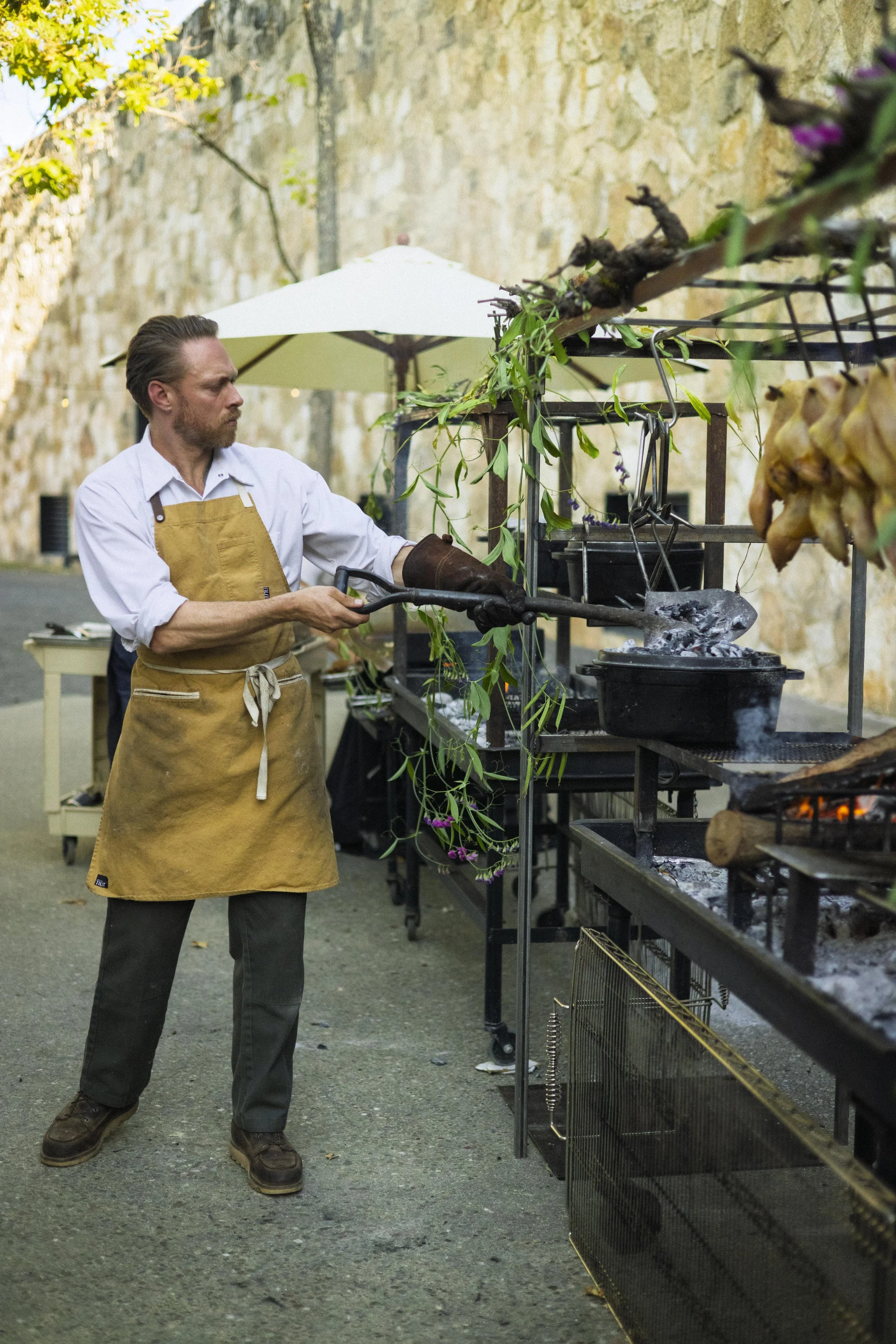 Man grilling food outdoors in a street setting, wearing a beige apron and gloves, next to a grill with flames and smoke, with a stone wall and patio umbrella in the background.