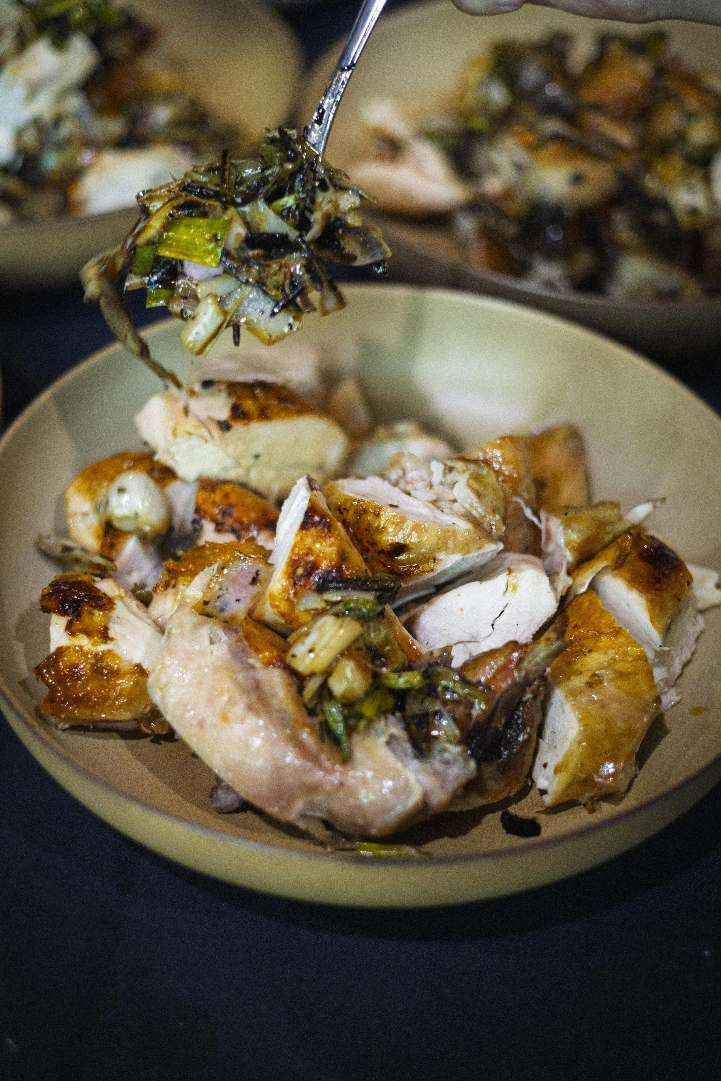 A bowl of roasted chicken topped with chopped green onions and served with side dishes in the background.