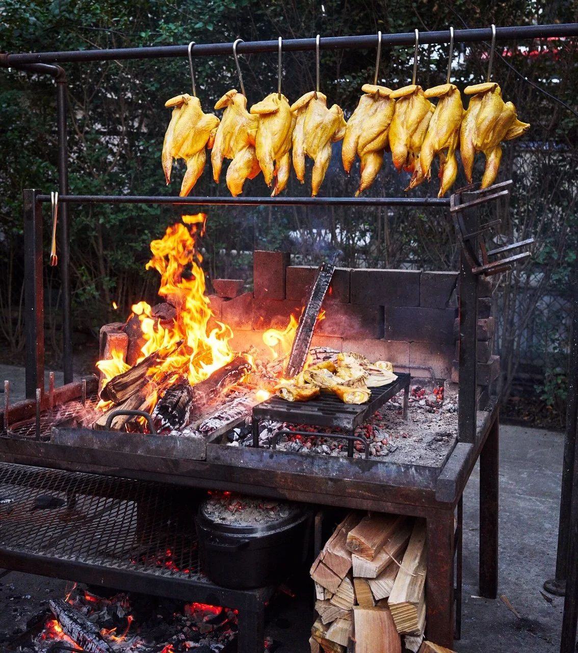 Chicken roasting on a rotisserie over an open flame, with some pieces cooking on a small grill nearby, and wood logs stored underneath the grill.