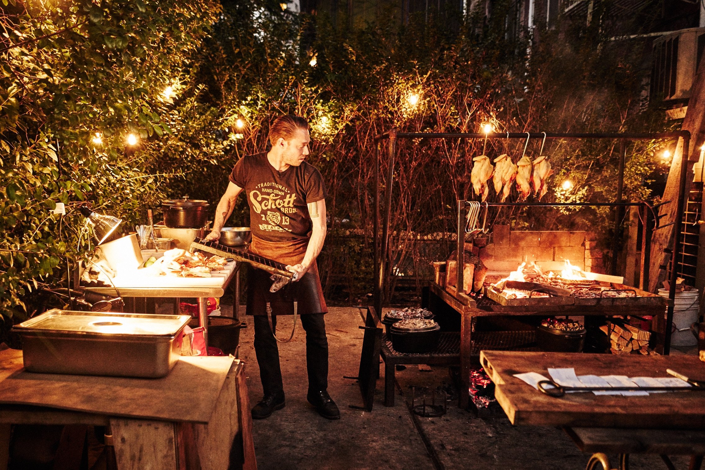 A man cooking outdoors at a backyard barbecue at night, with chicken on hooks above a fire pit and framed by string lights.