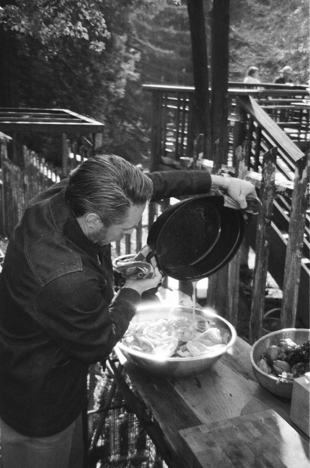A man in a jacket pouring sauce onto a bowl of food on an outdoor wooden picnic table with a fence and trees in the background.