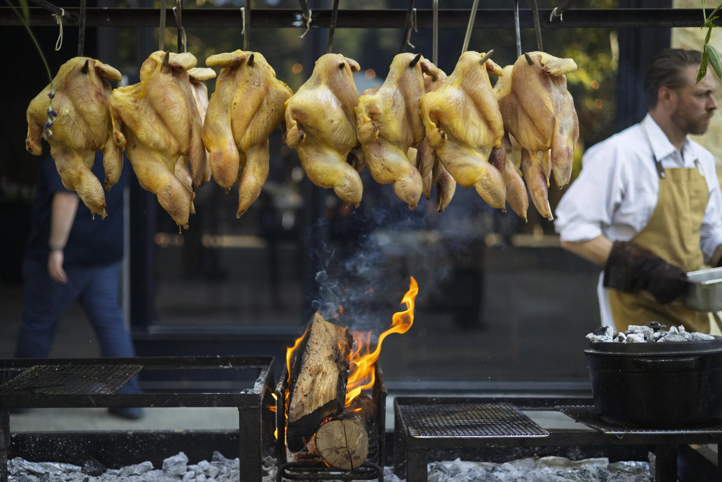 Hanging roasted chickens grilling over an open fire at an outdoor barbecue.
