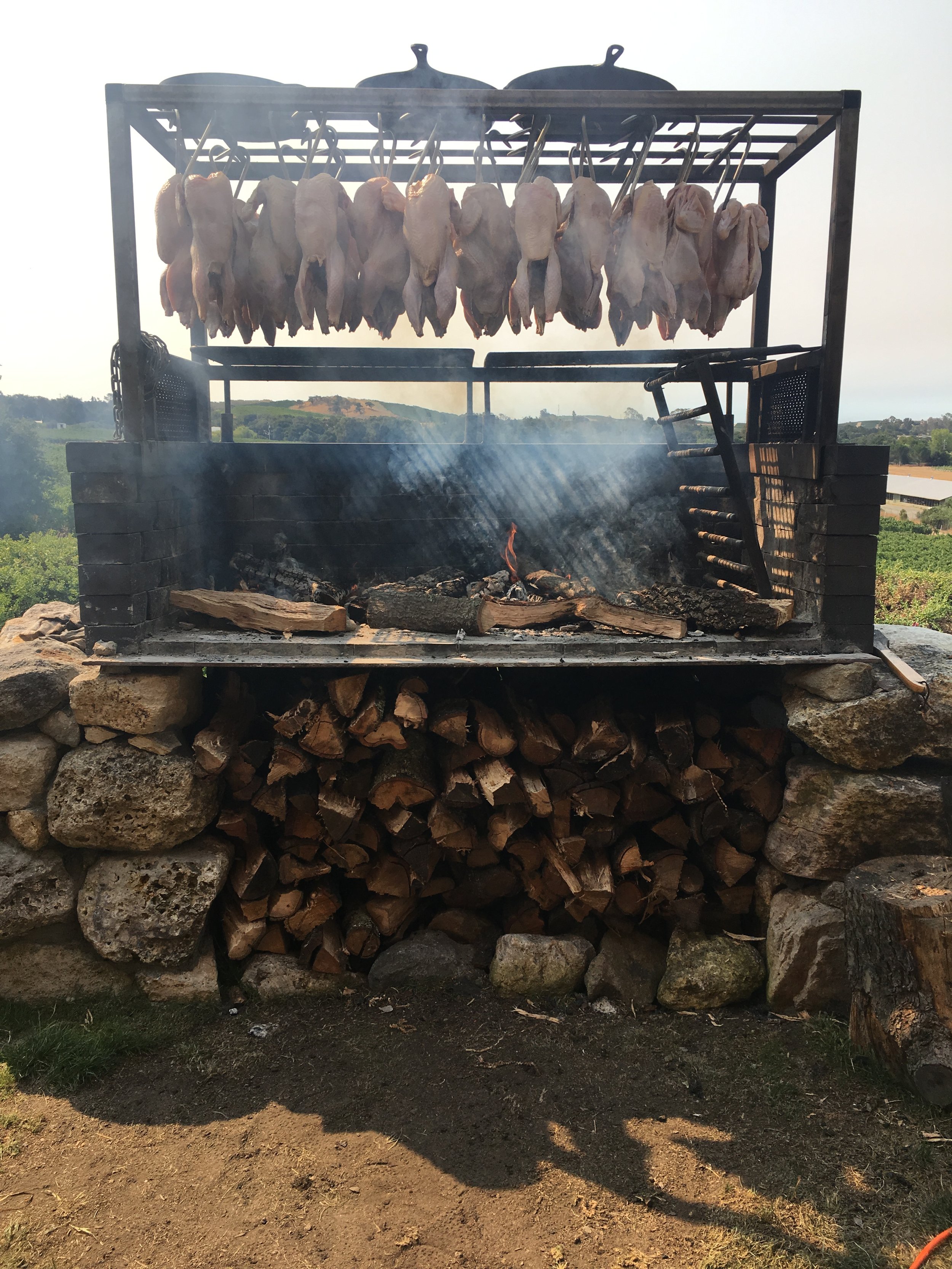 Chicken carcasses hanging on hooks over an outdoor wood-fired grill with stacked firewood underneath.