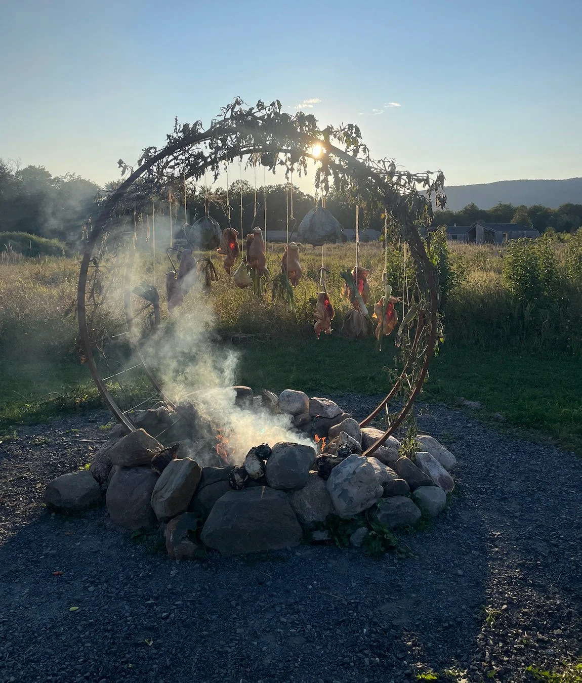 A rustic outdoor fire pit surrounded by rocks with a small fire burning and smoke rising, set in a rural field with a large arched metal frame overhead holding hanging dried gourds or vegetables, with trees, a building, and hills in the background at