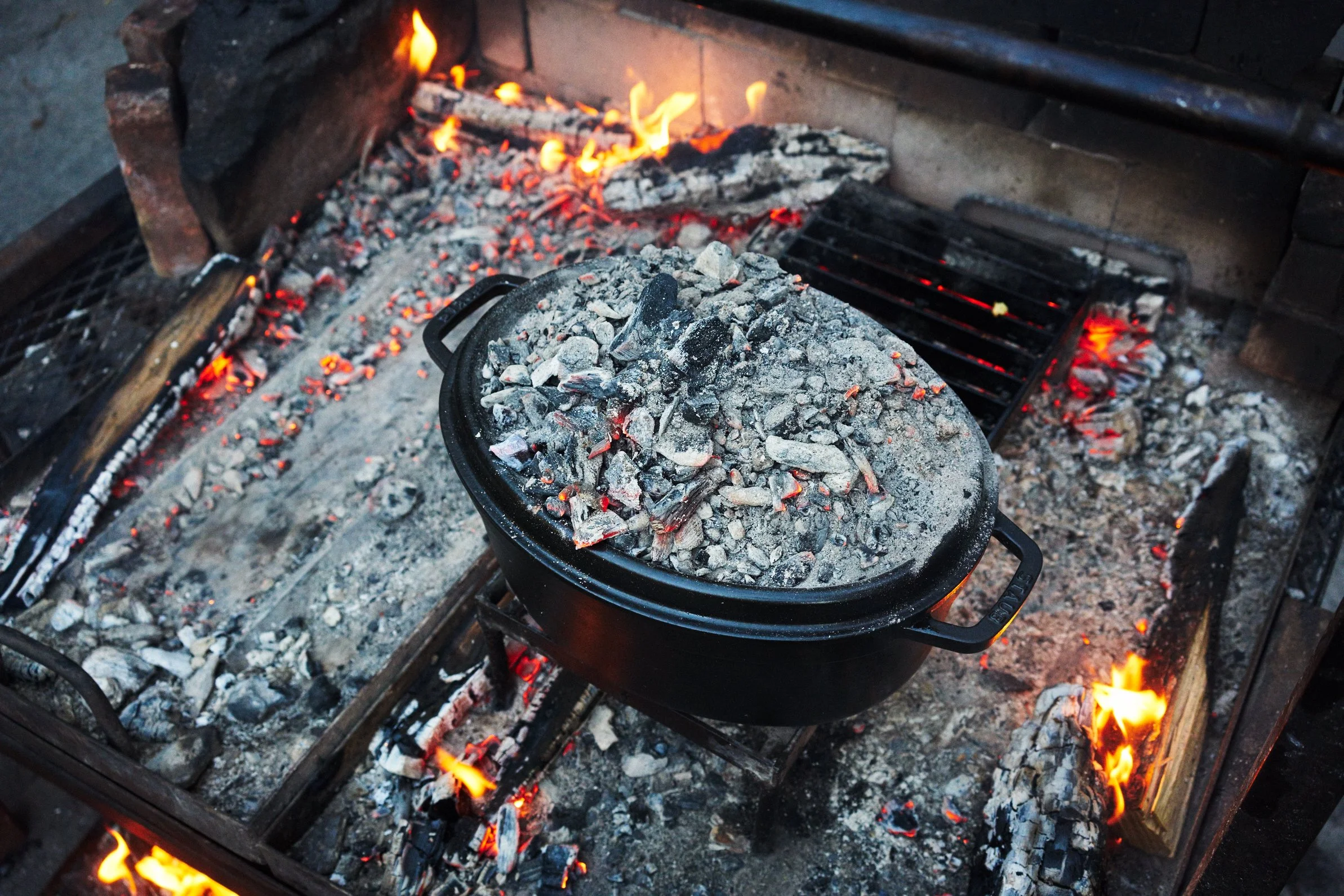 A black Dutch oven filled with hot charcoal sitting on a barbecue pit with burning wood and glowing coals.