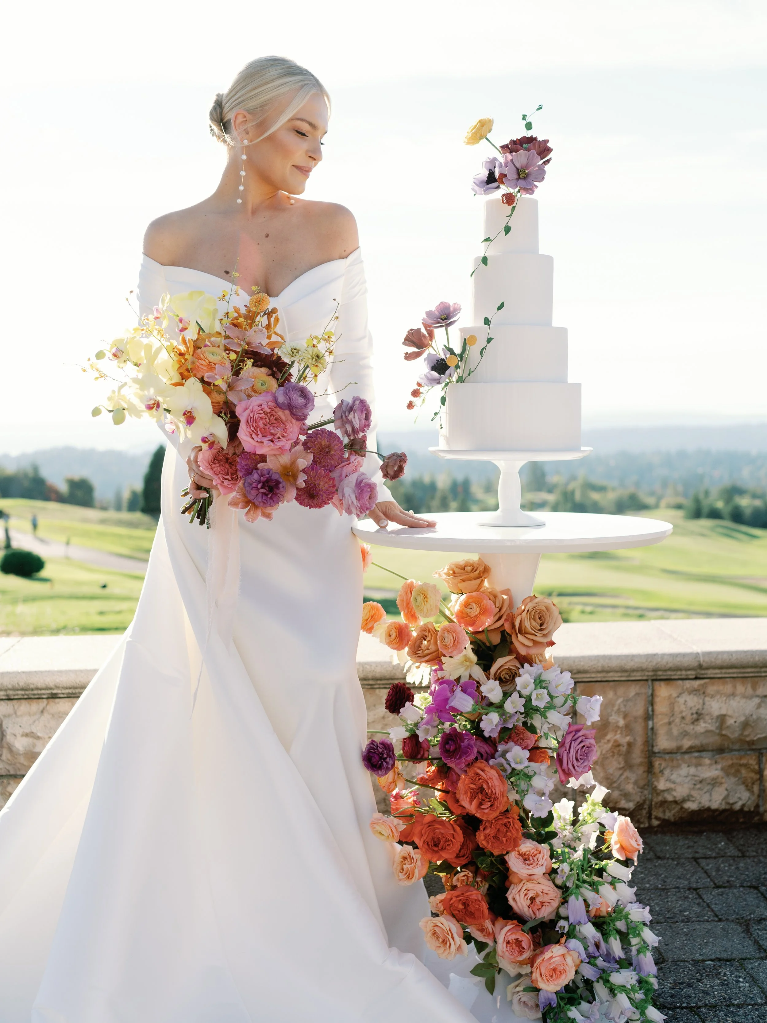 A bride in a white wedding dress holding a large bouquet of colorful flowers, standing next to a tall white tiered wedding cake decorated with flowers, outdoors with a scenic background of green hills and a bright sky.