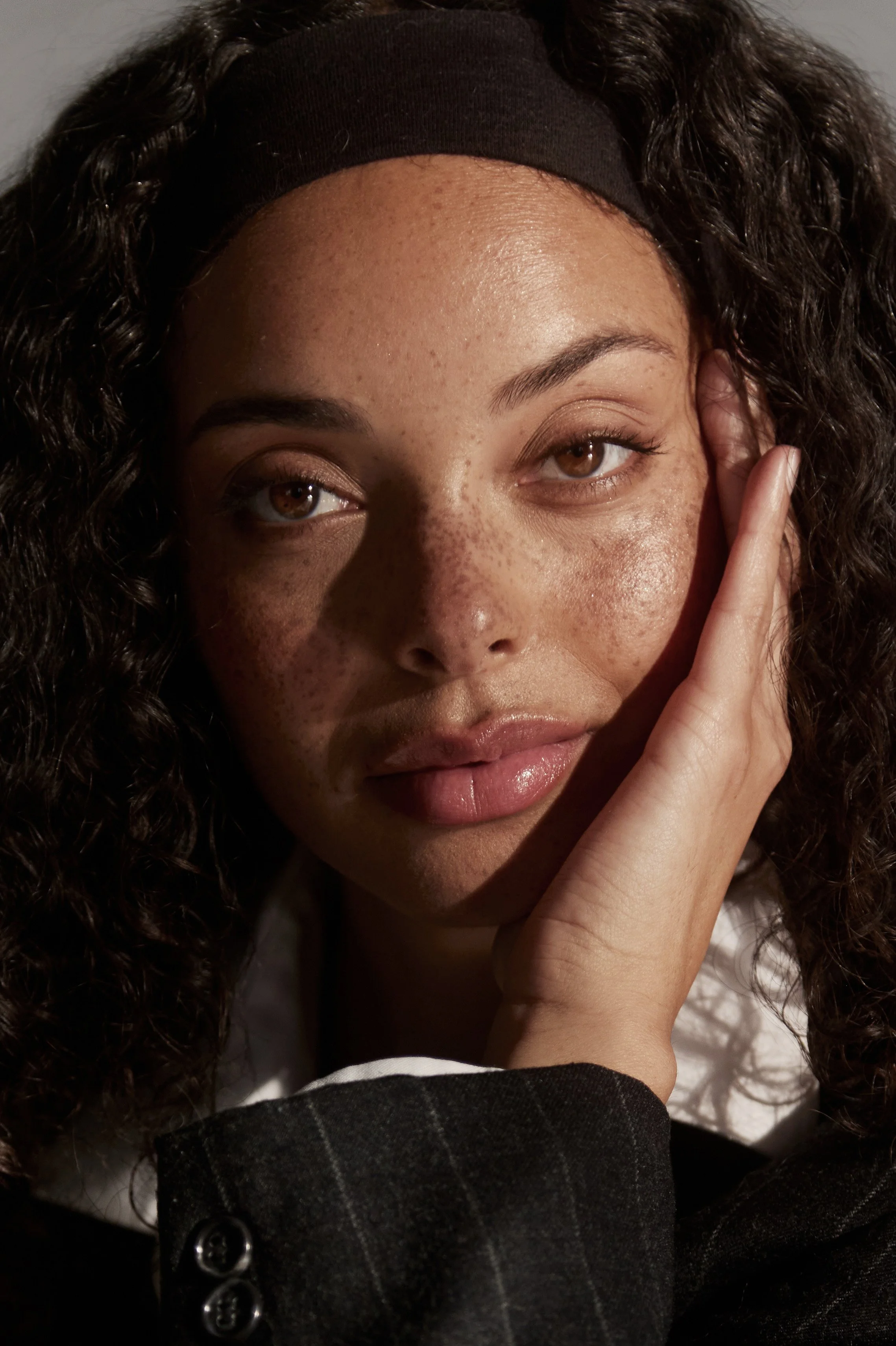 A close-up of a woman with curly hair, wearing a black headband and black blazer, touching her face with her hand.
