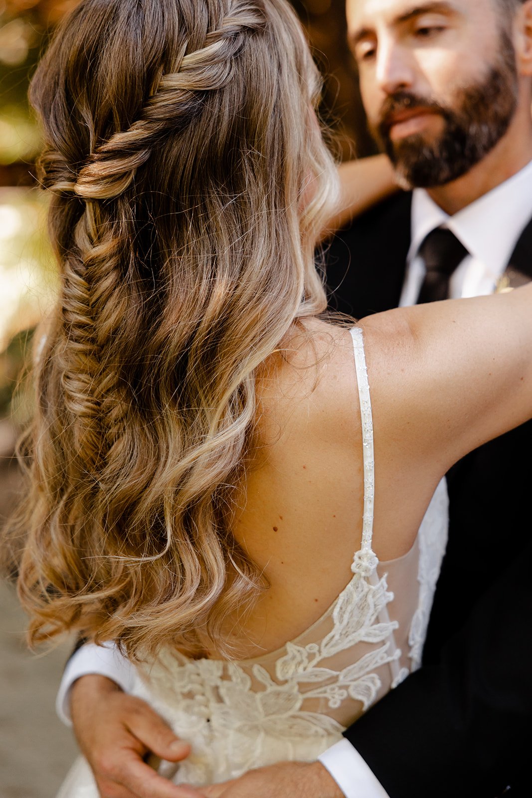 A bride and groom are dancing closely, with the bride's long, wavy hair and lace dress visible, and the groom wearing a black tuxedo and tie.