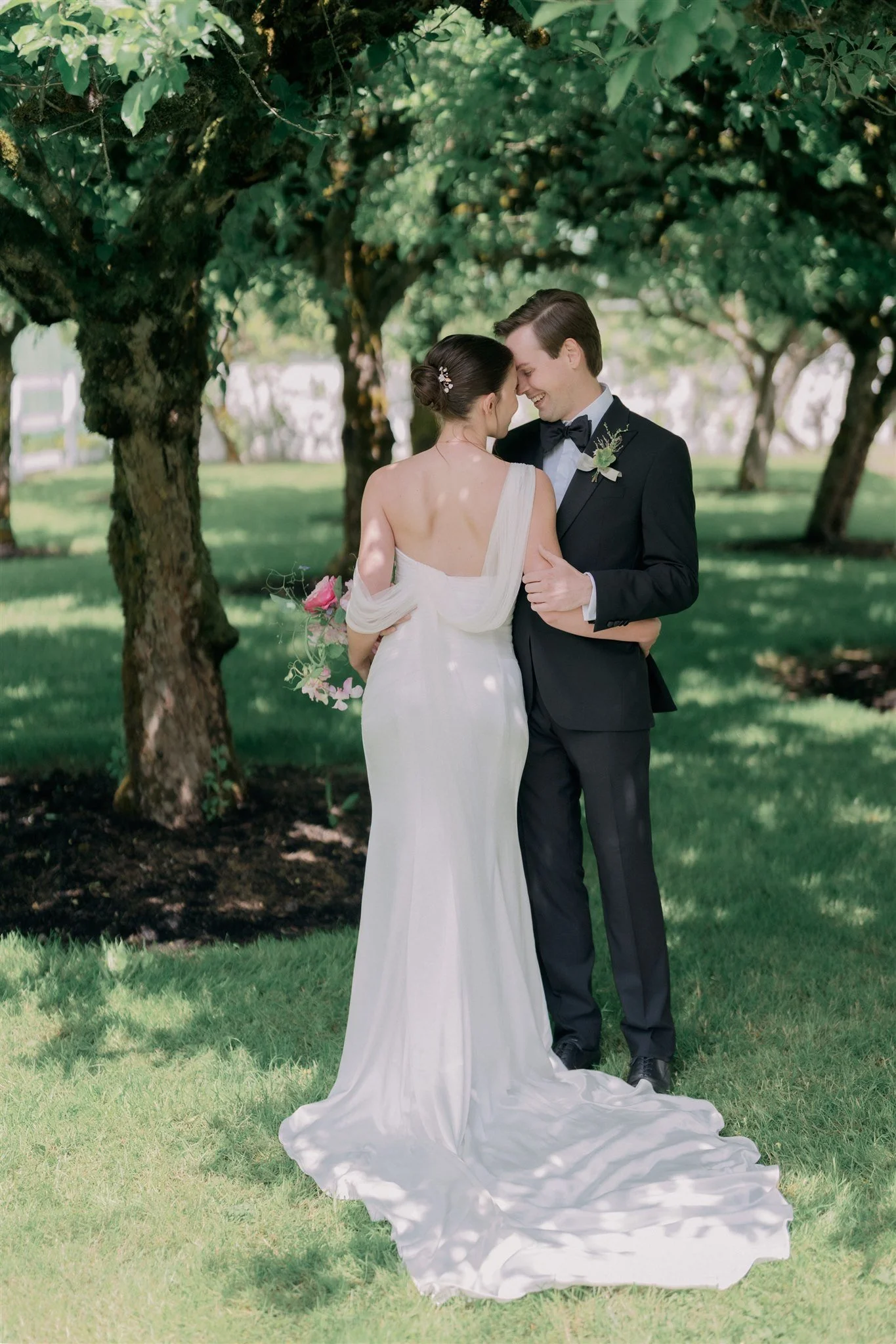 A bride and groom sharing a moment on their wedding day outdoors, standing closely under trees with lush greenery.