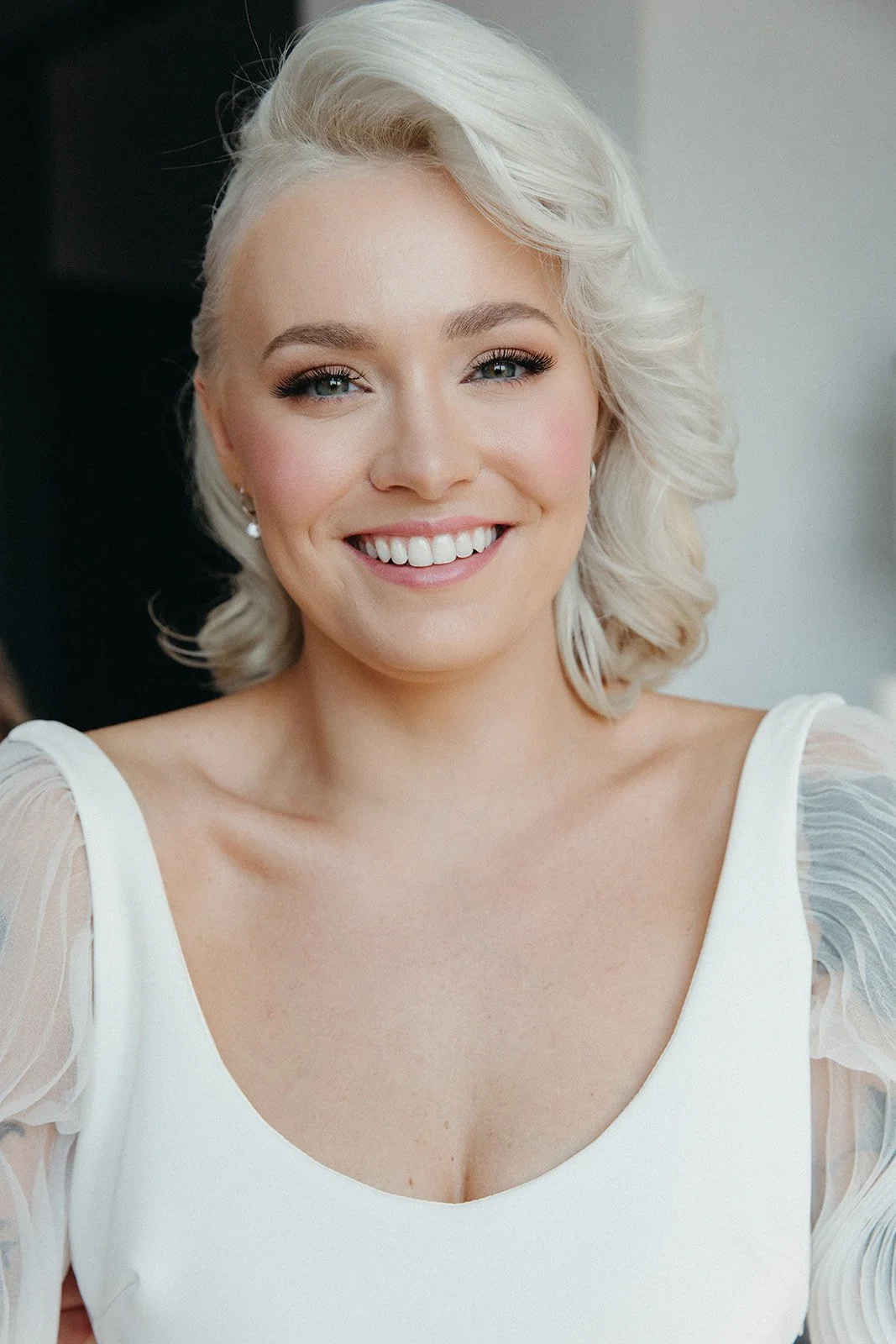 Close-up of a smiling woman with light blonde, wavy hair and blue eyes, wearing a white dress with sheer puff sleeves.