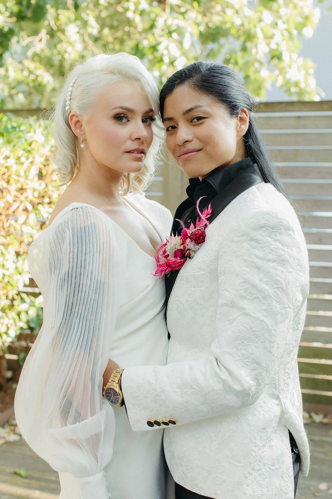 Two women, one with platinum blonde hair and the other with black hair, dressed in white and black formal suits, standing closely together outdoors with a wooden fence and greenery in the background.