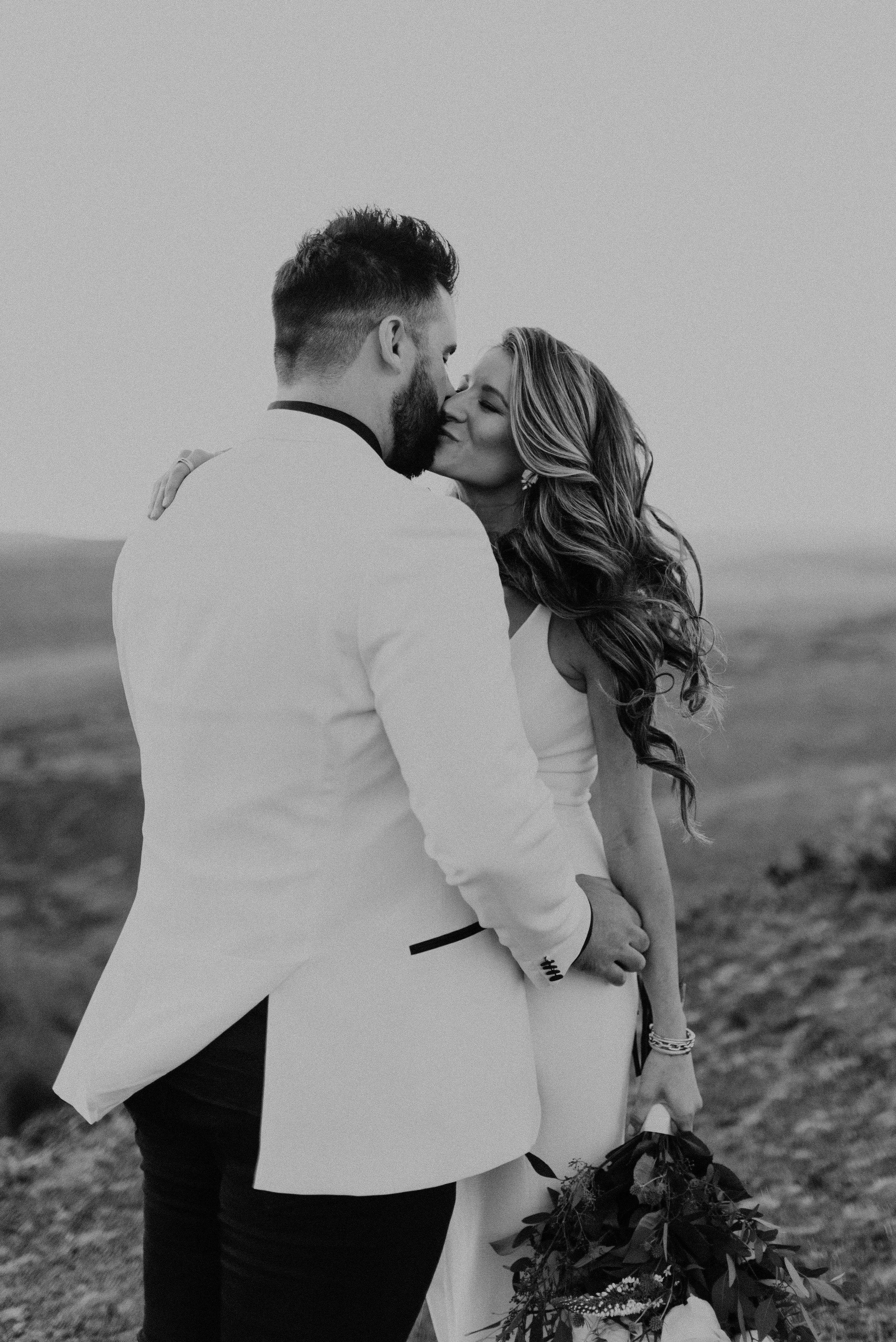A black and white photo of a couple dressed formally, embracing each other outdoors, with the woman holding a bouquet of flowers.