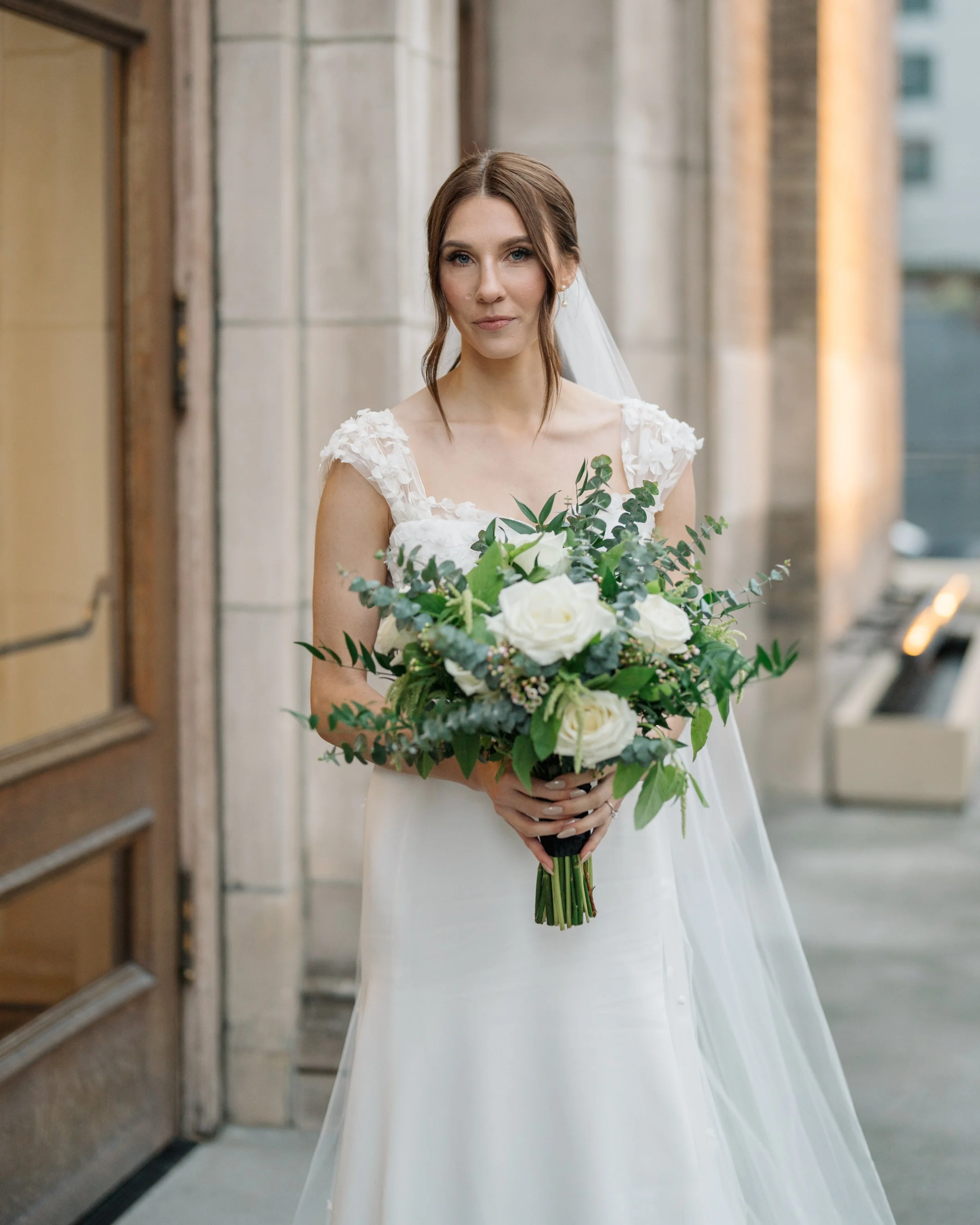 A bride wearing a white wedding dress holding a bouquet of white roses and green foliage, standing outside near a building with stone and wooden elements.