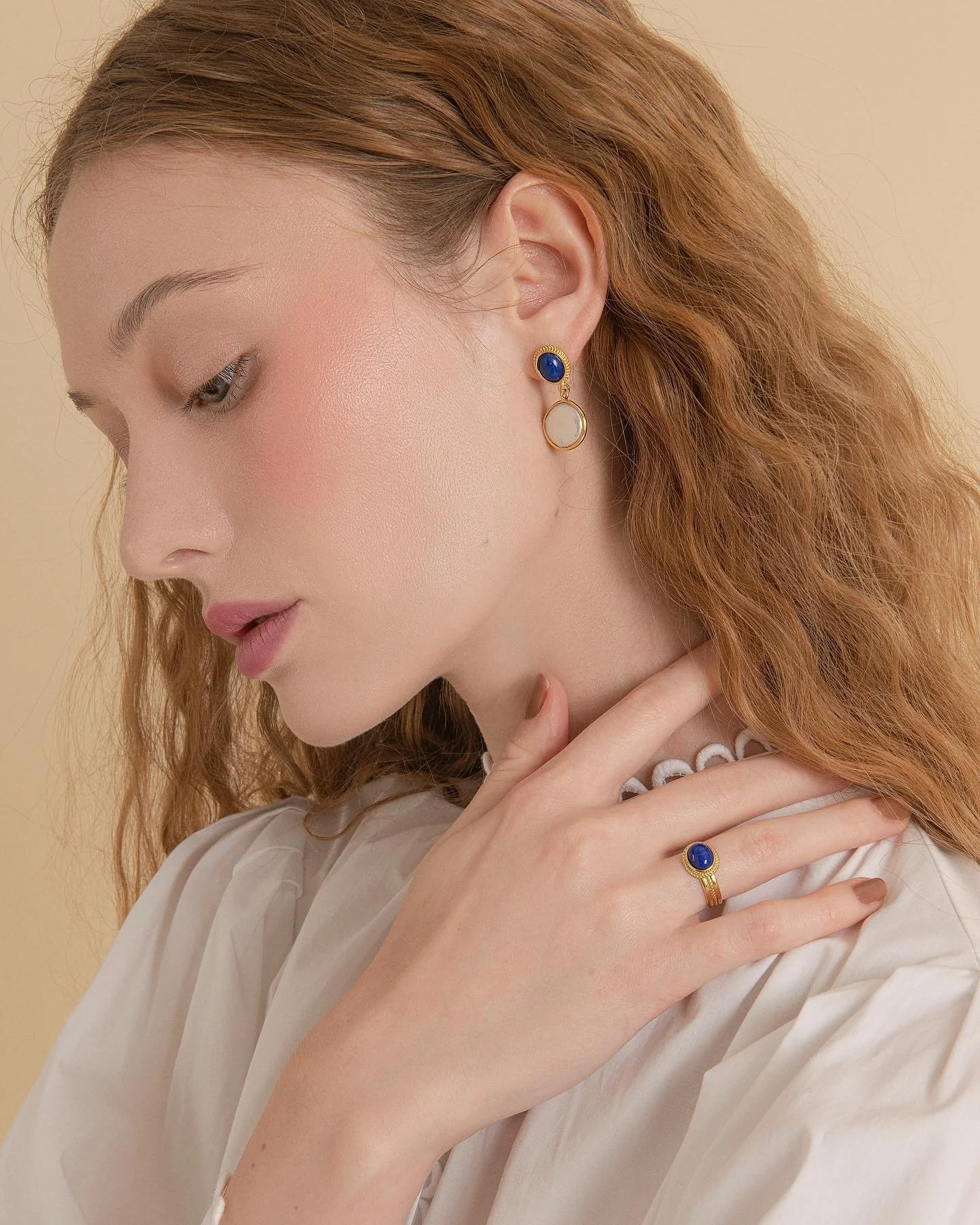 Close-up of a woman with red hair wearing blue and white jewelry, including earrings, a ring, and a necklace, against a beige background.