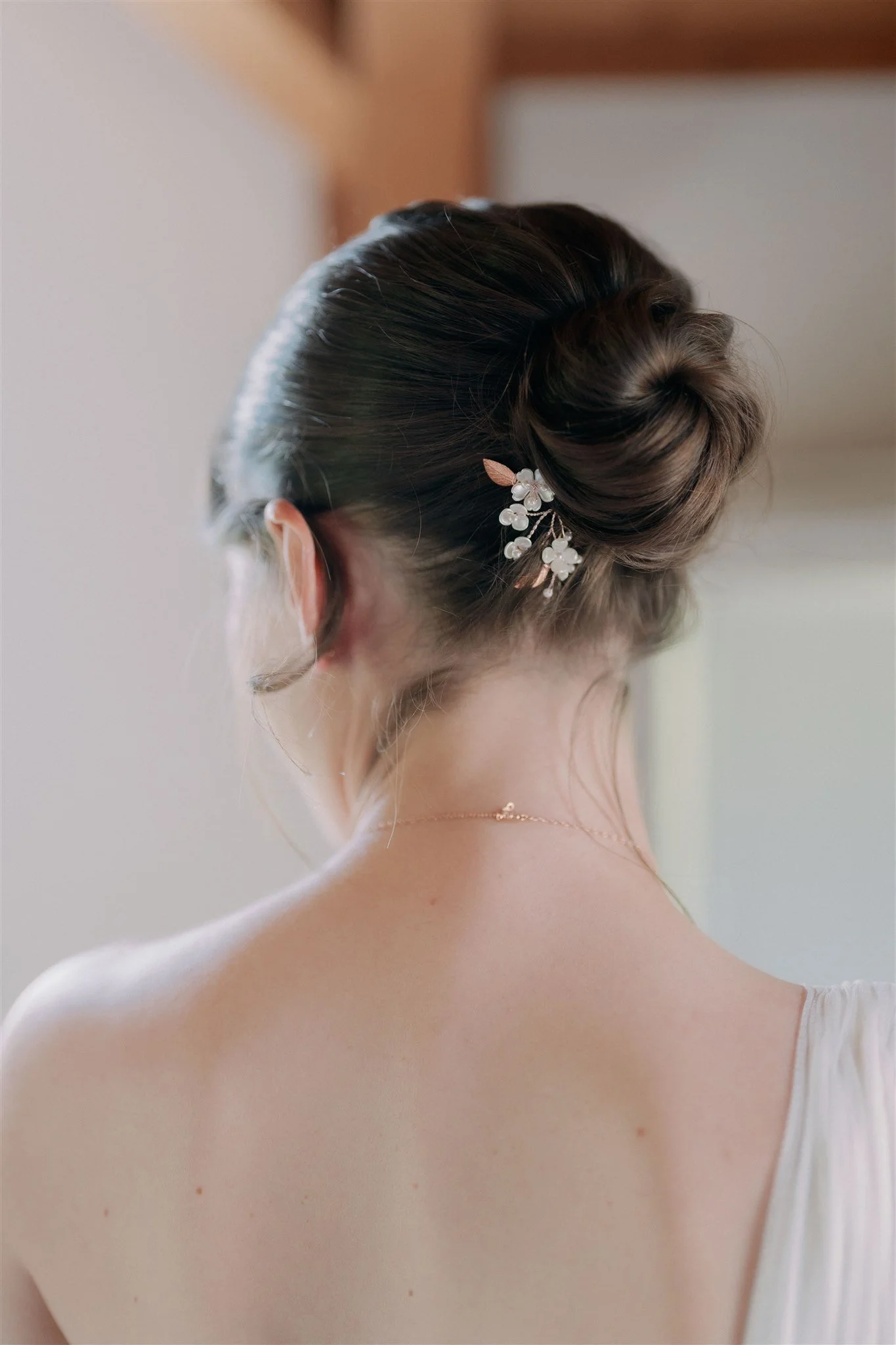 A woman with dark hair styled in an updo adorned with a pearl and leaf hairpiece, shown from behind, wearing a white dress.