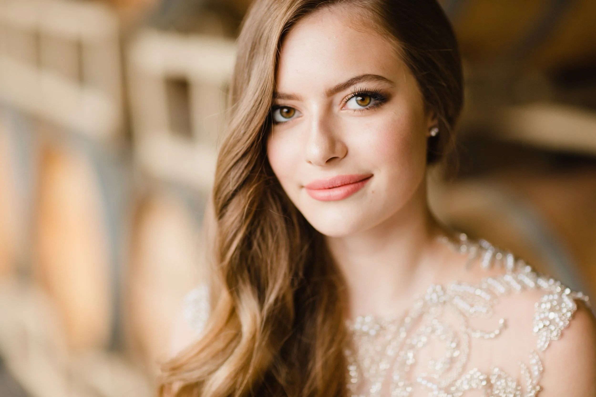 Close-up of a young woman with long, wavy brown hair, wearing makeup with pink lipstick and jewelry, looking at the camera with a slight smile.