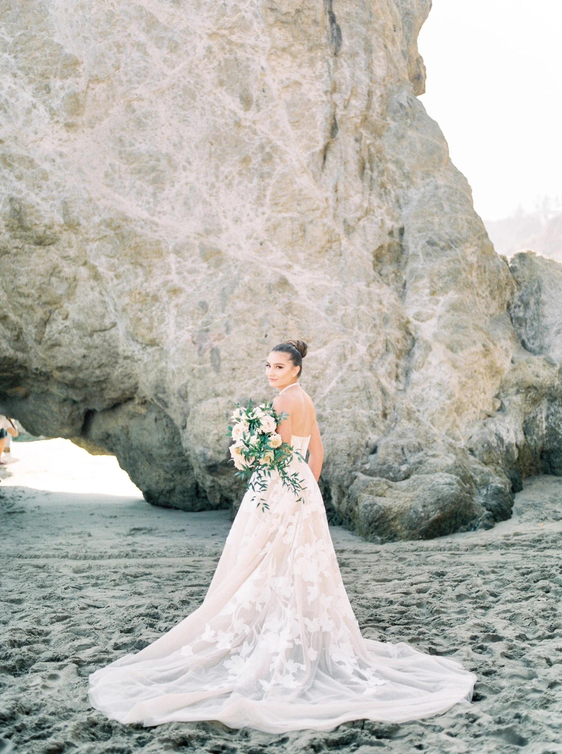 A woman in a wedding dress holding a bouquet, standing on a sandy beach with large rock formations in the background.