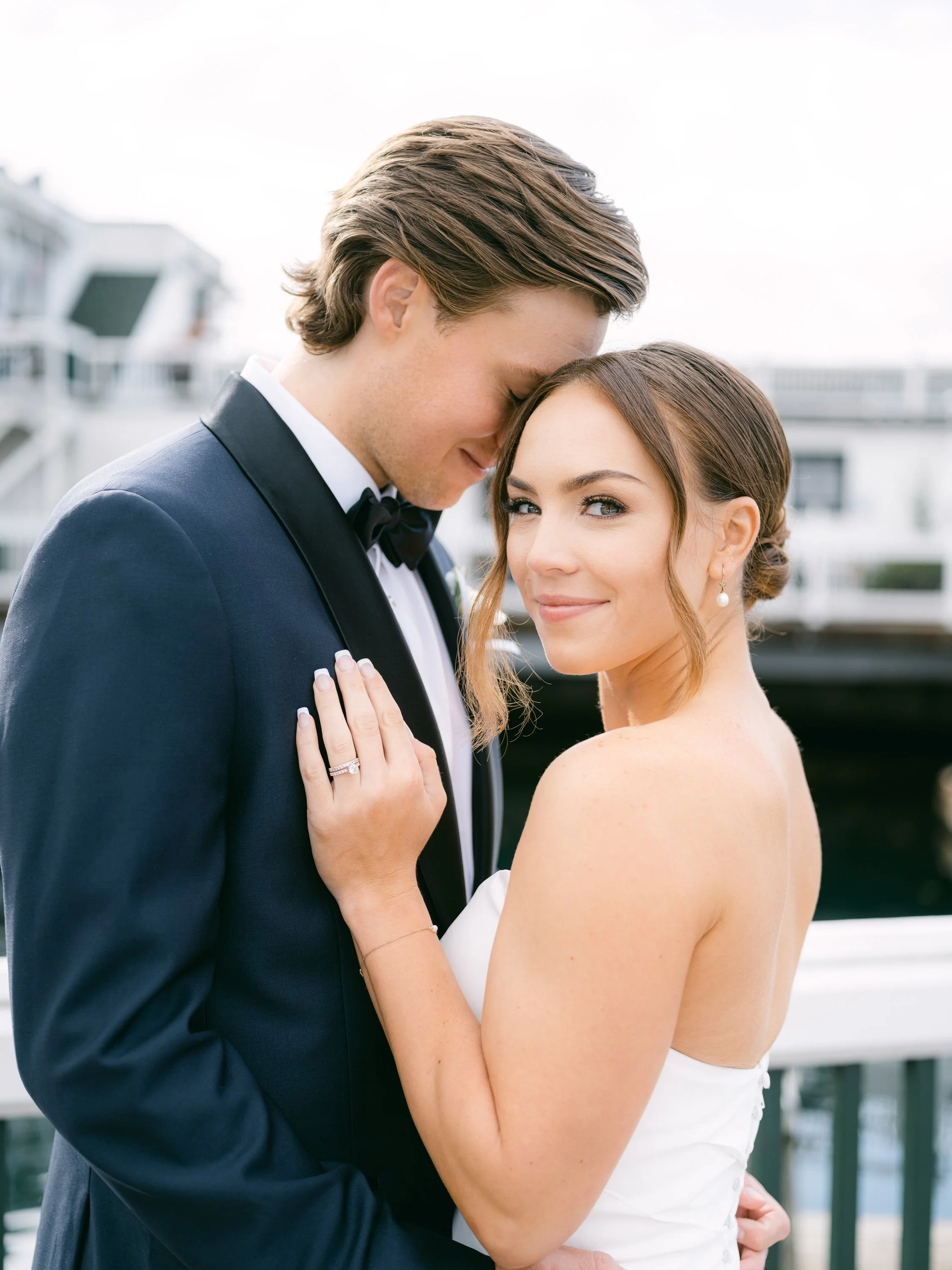A bride and groom on their wedding day standing close together outdoors, with the groom resting his forehead against the bride's forehead and the bride smiling softly, wearing pearl earrings and a strapless wedding dress.