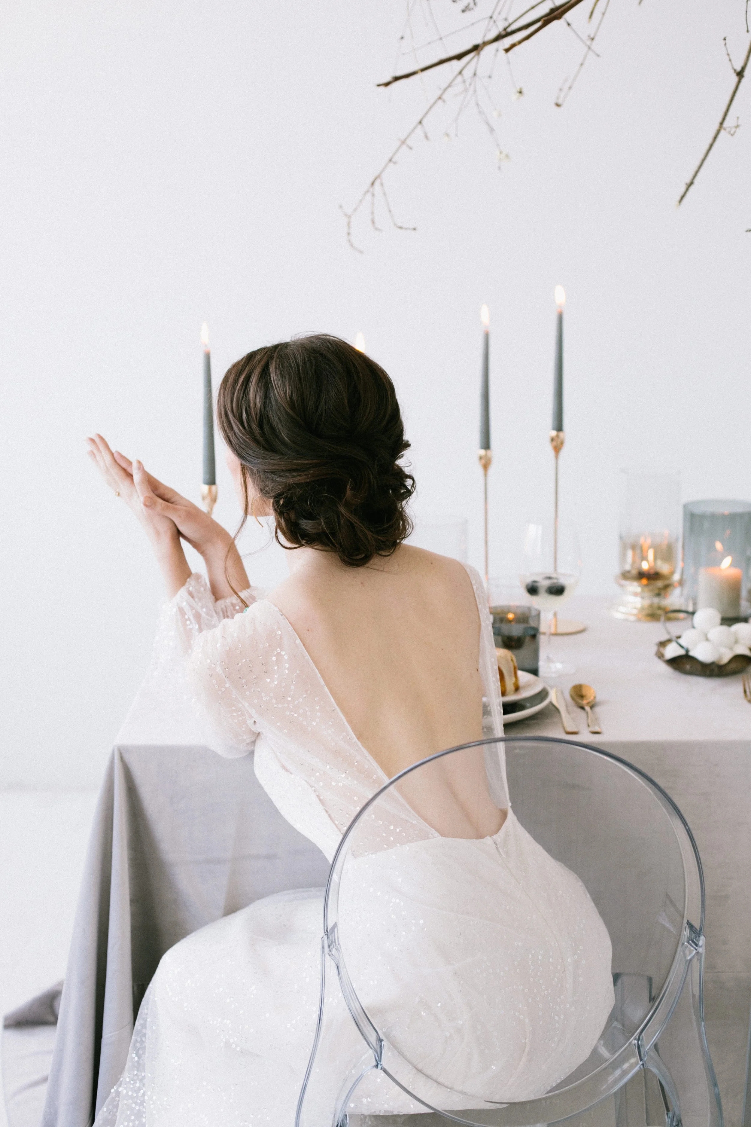 A woman with brown hair styled in soft waves, seen from the back, sitting at a table with a beige tablecloth and a semi-transparent acrylic chair. The woman wears a white, off-shoulder dress with delicate sparkle embellishments. In the background, th