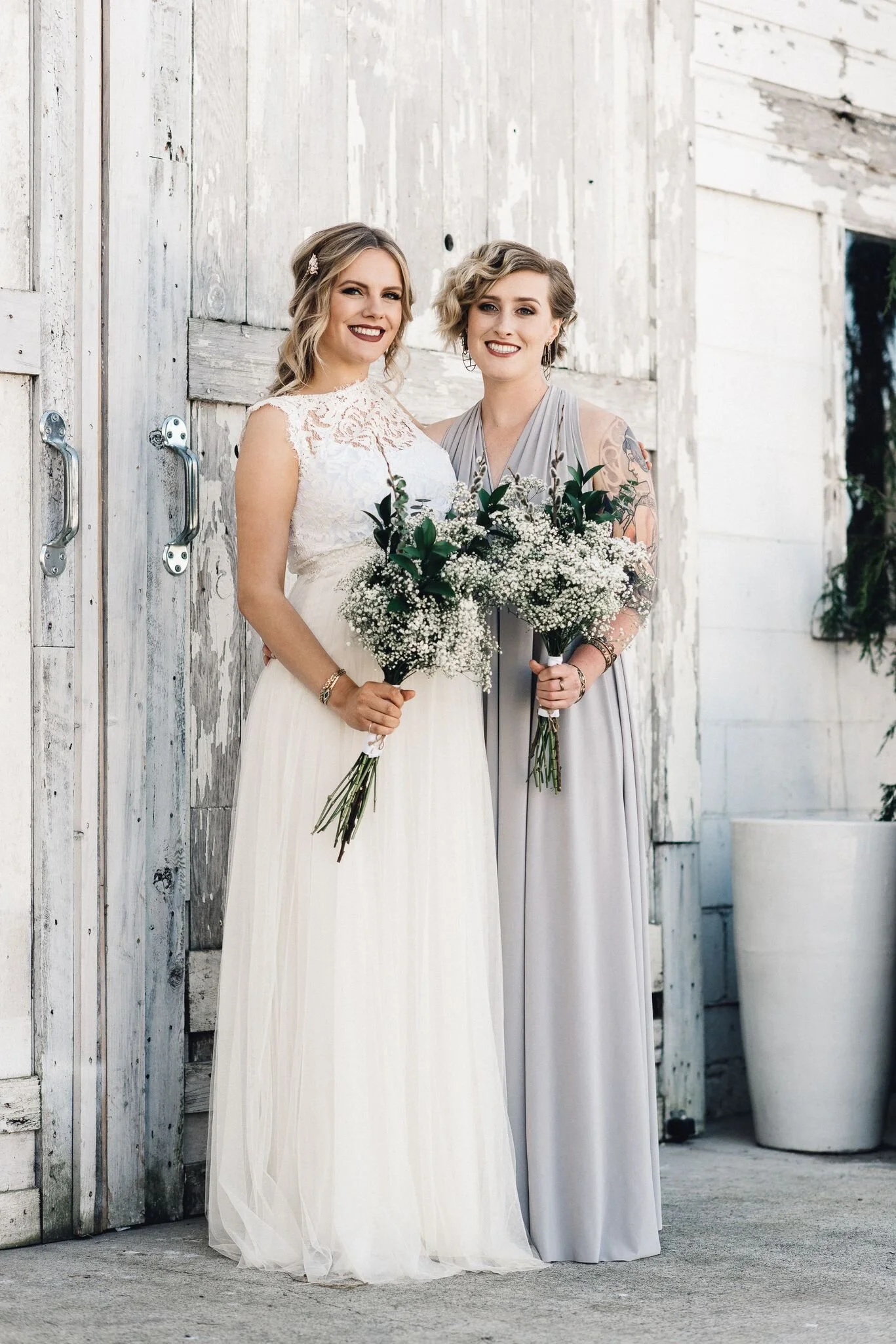 Two women in wedding attire holding bouquets, standing in front of a rustic white wooden backdrop.
