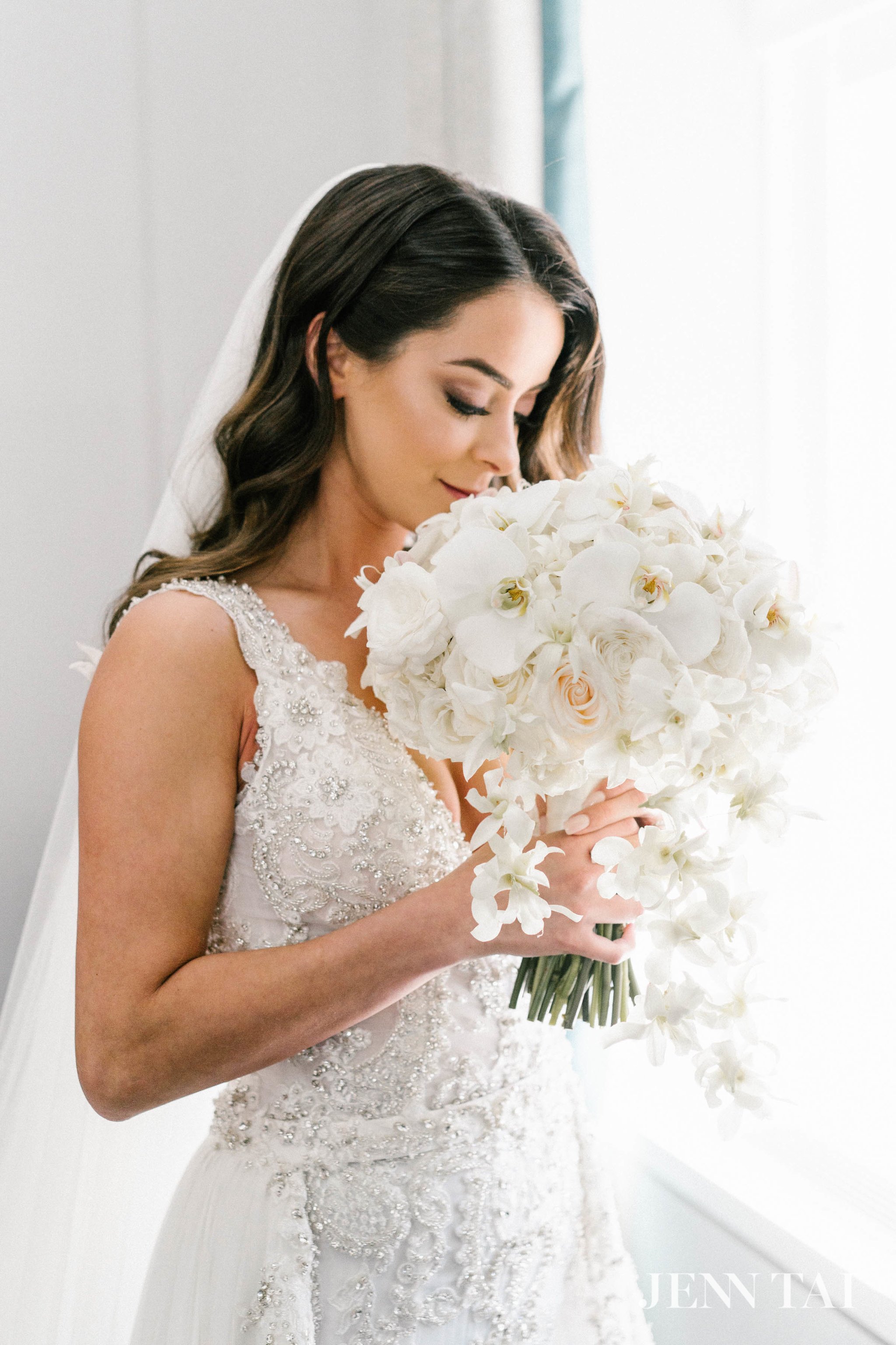 A bride in a white wedding dress holding a large bouquet of white orchids and roses, with long dark hair styled in loose waves and a light veil.