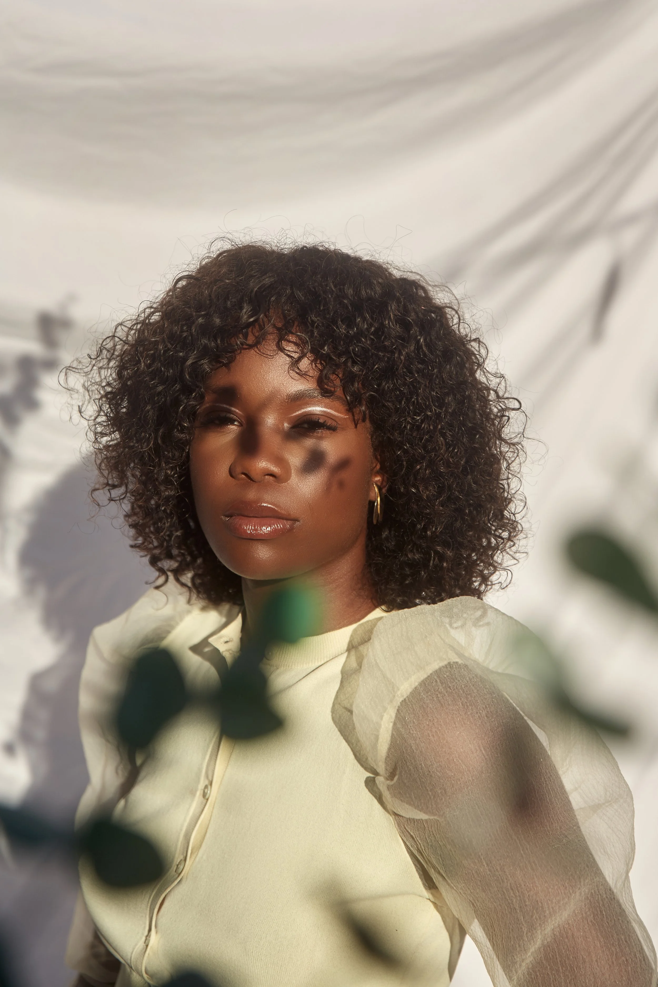 Close-up of a woman with curly hair and gold earrings, wearing a light yellow blouse with sheer, puffed sleeves, standing against a white cloudy background with shadows.