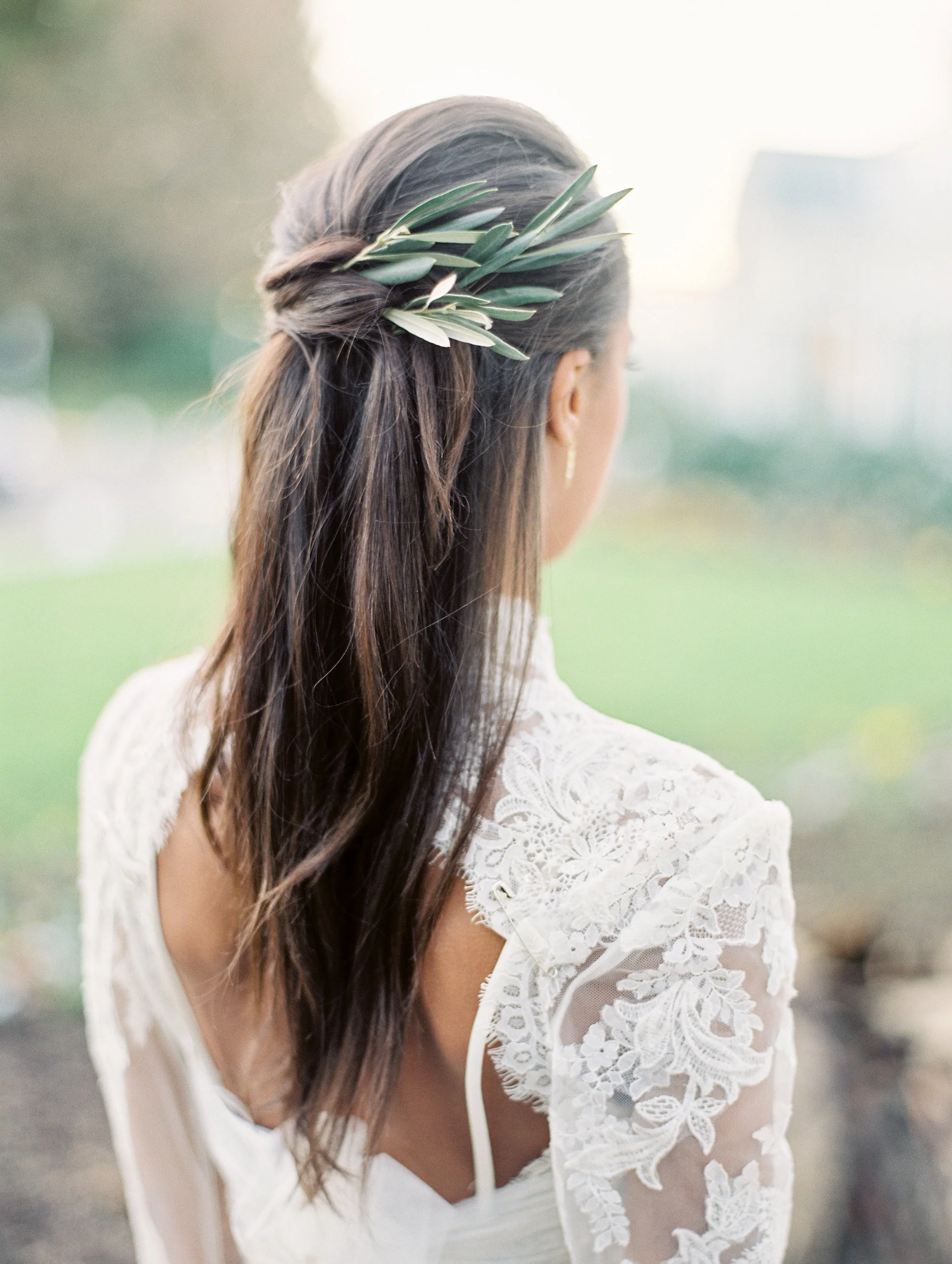 A woman with long brown hair styled half-up, wearing a white lace dress and a green olive branch hair accessory, standing outdoors during daytime.