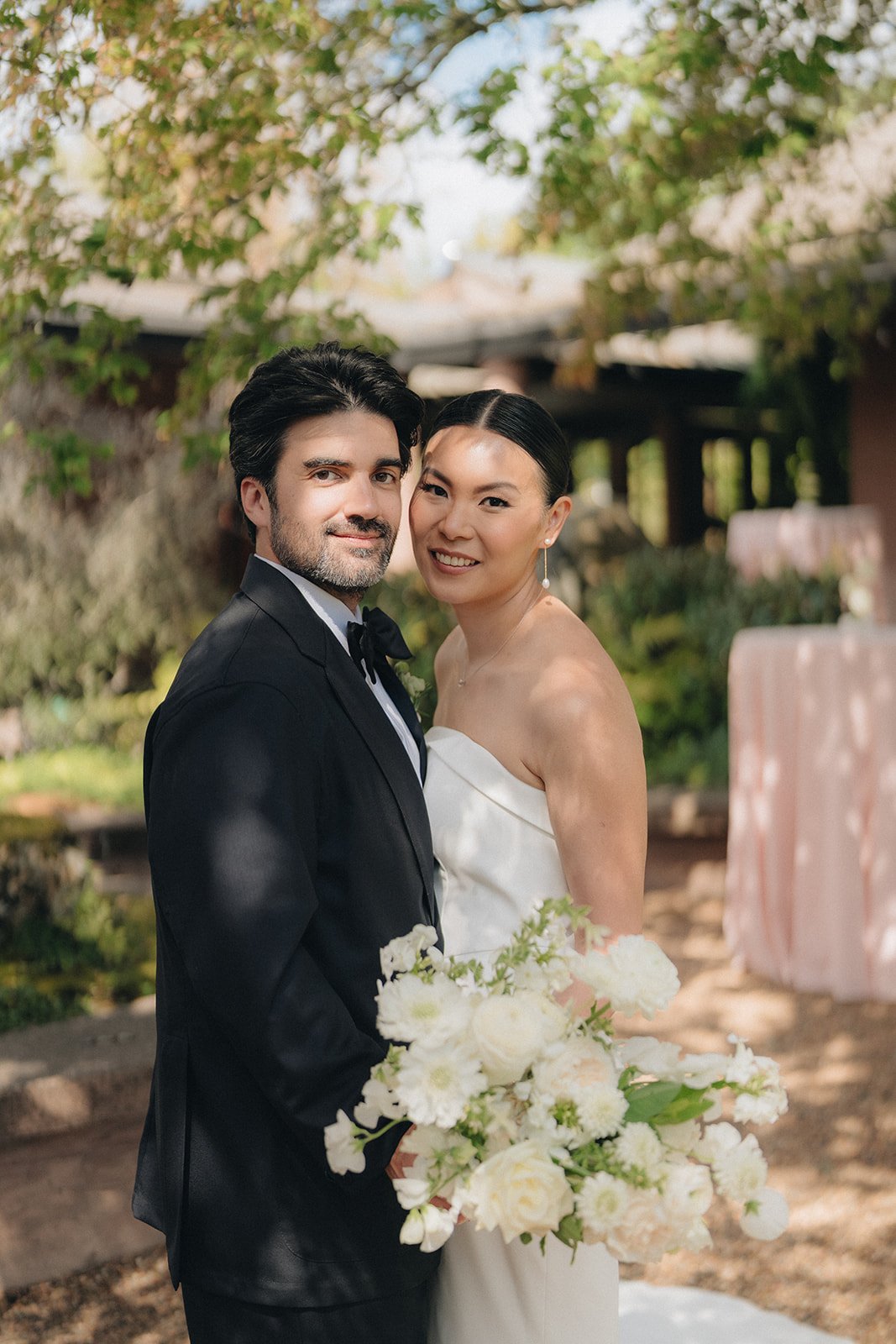 A newlywed couple standing outdoors, smiling at the camera, with the bride holding a large bouquet of white flowers, surrounded by green trees and outdoor decor.
Karla makeup, Meghann hair
Photographer Kris Ladera 
Planner Lyndsay A Renee weddings
Ve