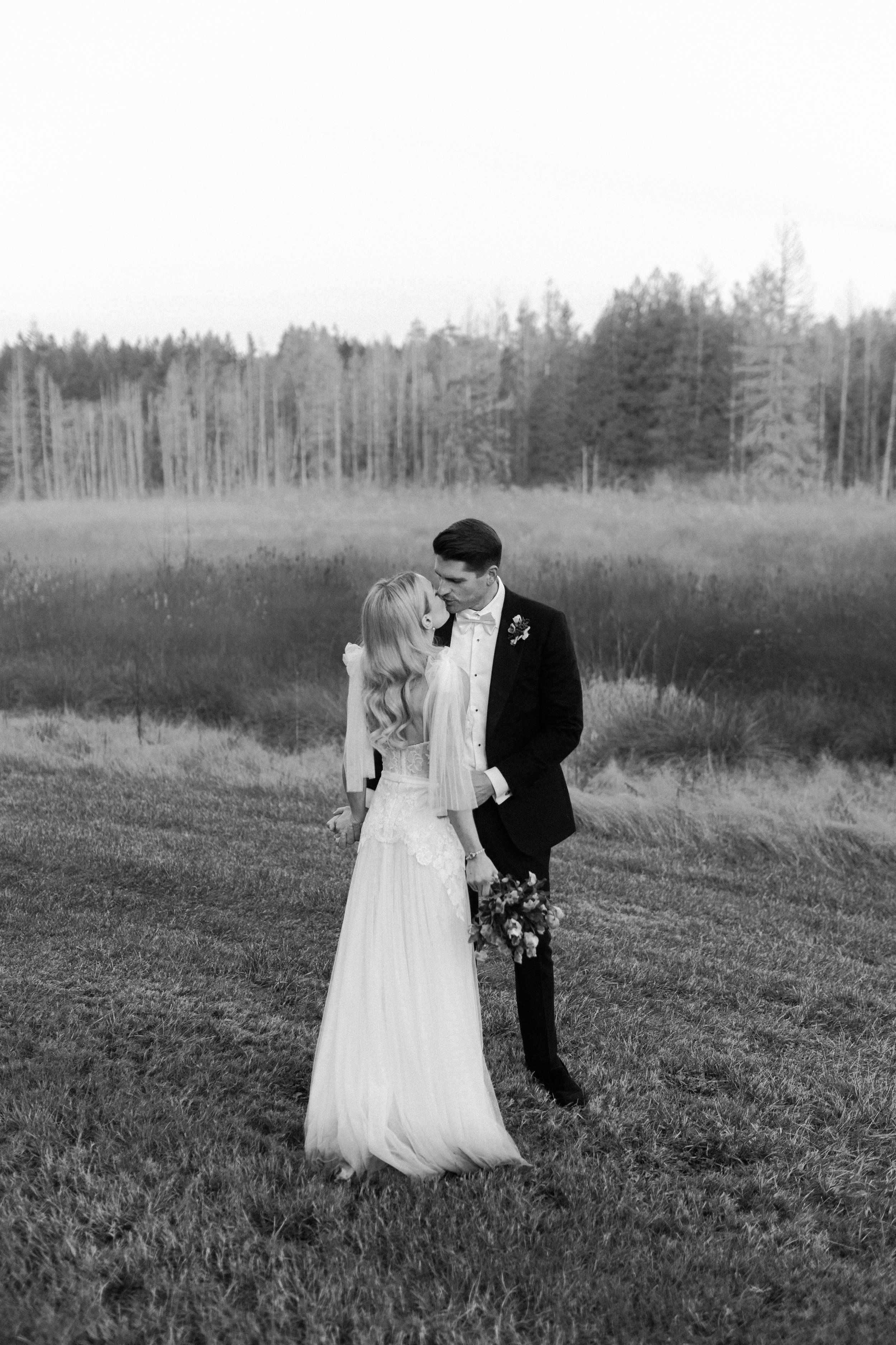 A black and white photograph of a bride and groom standing in a grassy field, with the bride holding a bouquet of flowers and the groom in a tuxedo. They are sharing an intimate moment, looking into each other's eyes, with a forest in the background.