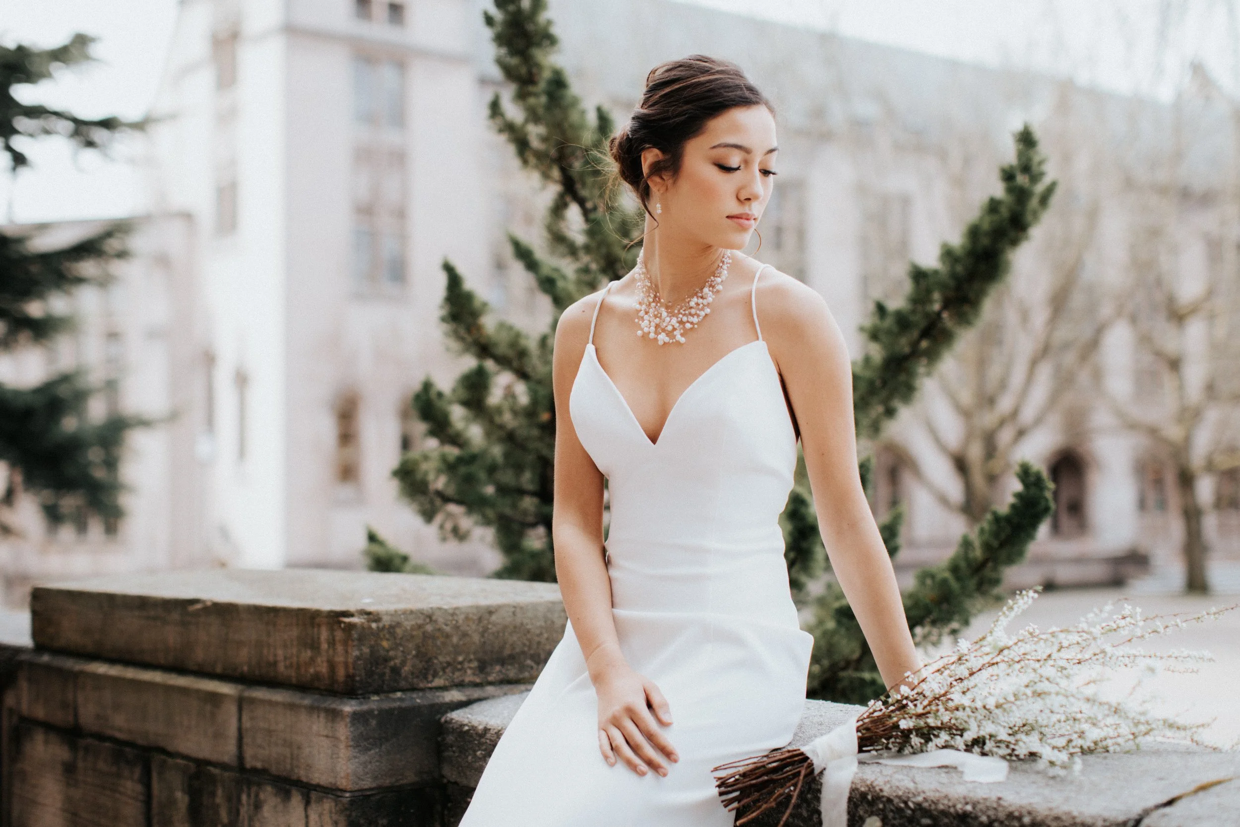 A woman in a white wedding dress standing outdoors with trees and buildings in the background, holding a bouquet of floral branches.