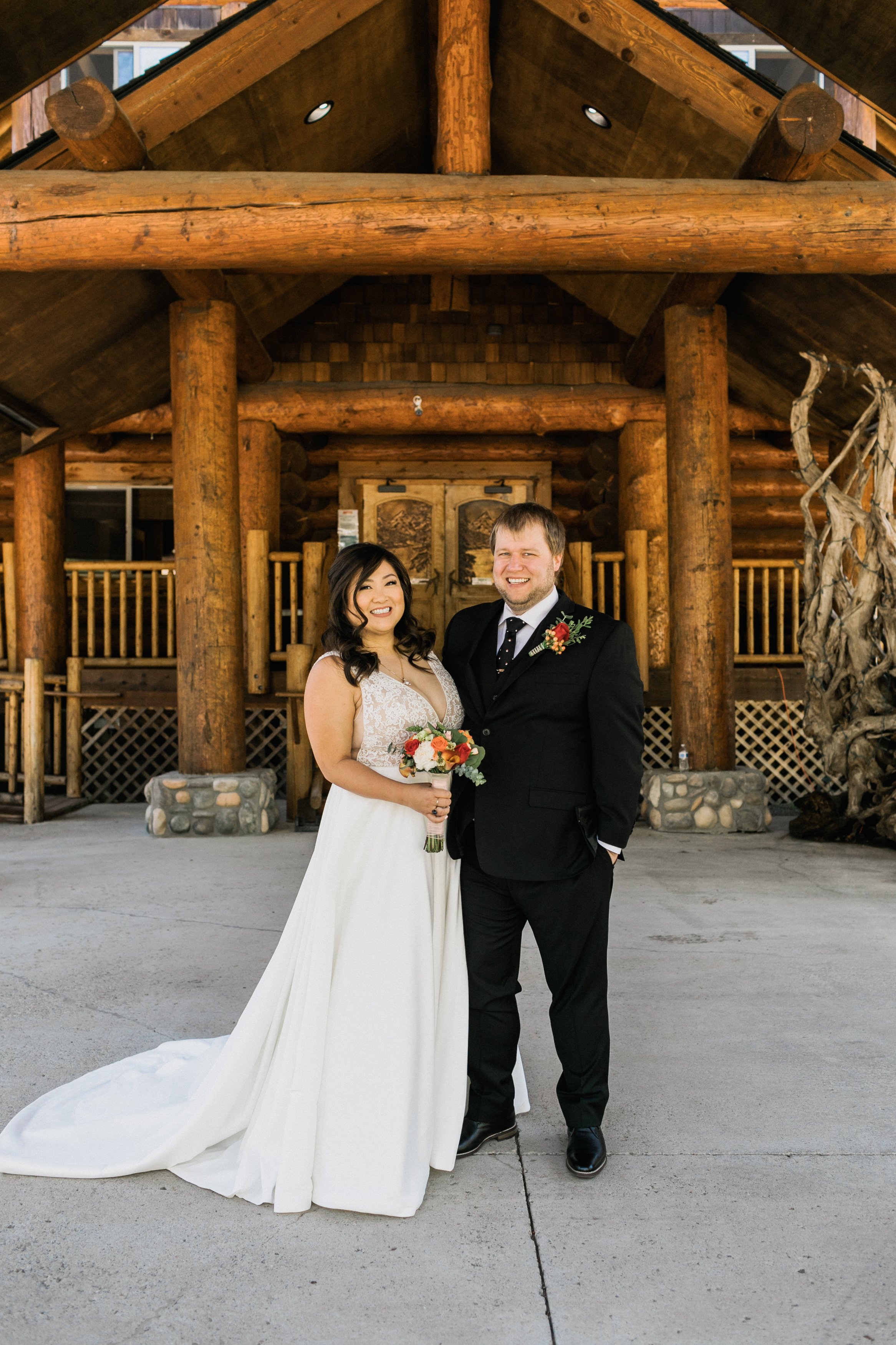 A bride and groom standing together outdoors in front of a wooden lodge, smiling and posing for a wedding photo. The bride is holding a bouquet of flowers, wearing a white dress, and the groom is in a black suit with a tie, both appearing happy and j