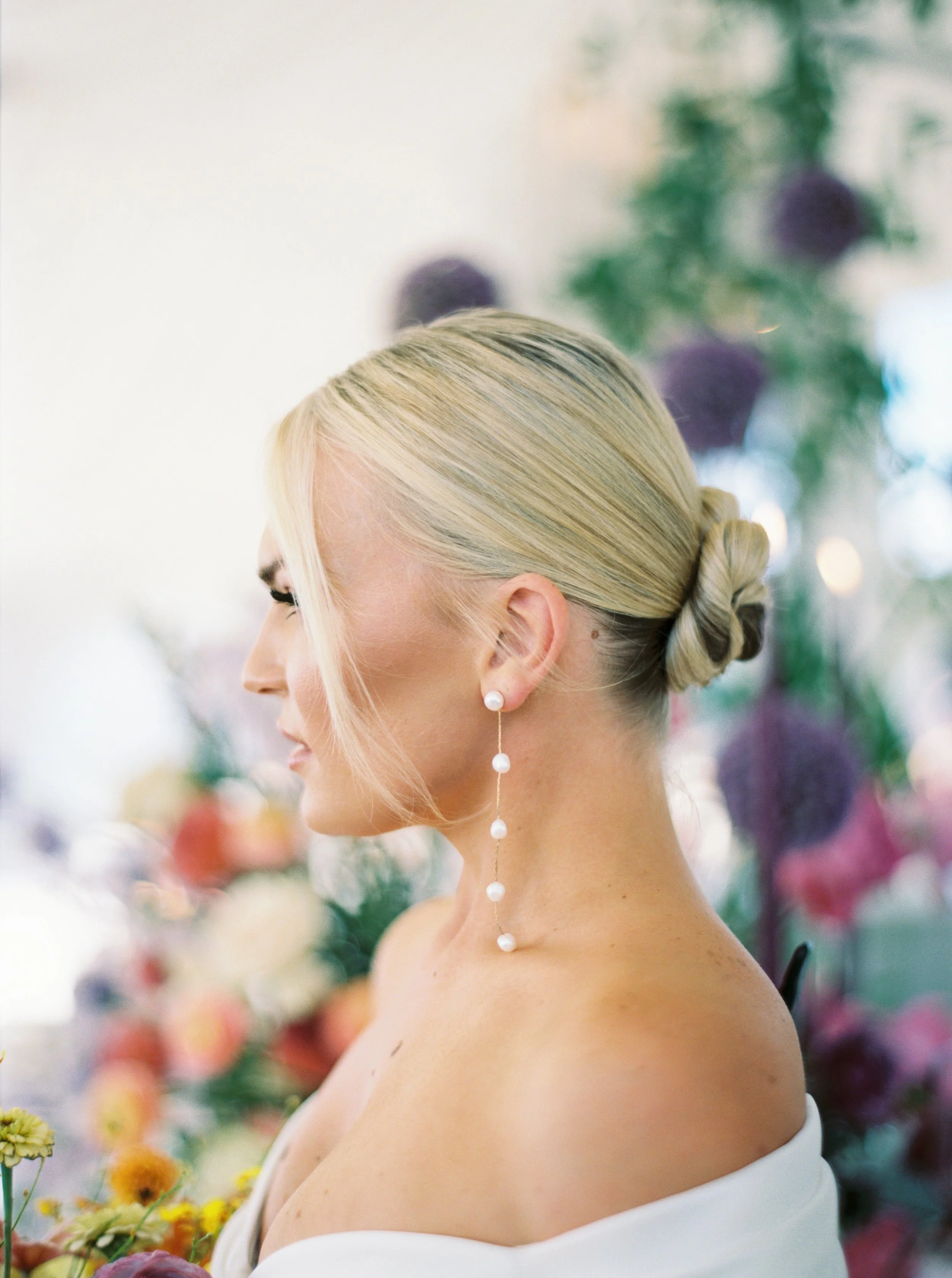 A woman with blonde hair styled in an elegant updo with a twisted bun, wearing long pearl earrings and a white off-shoulder dress, is surrounded by a blurred background of colorful flowers, possibly at a wedding or special event.