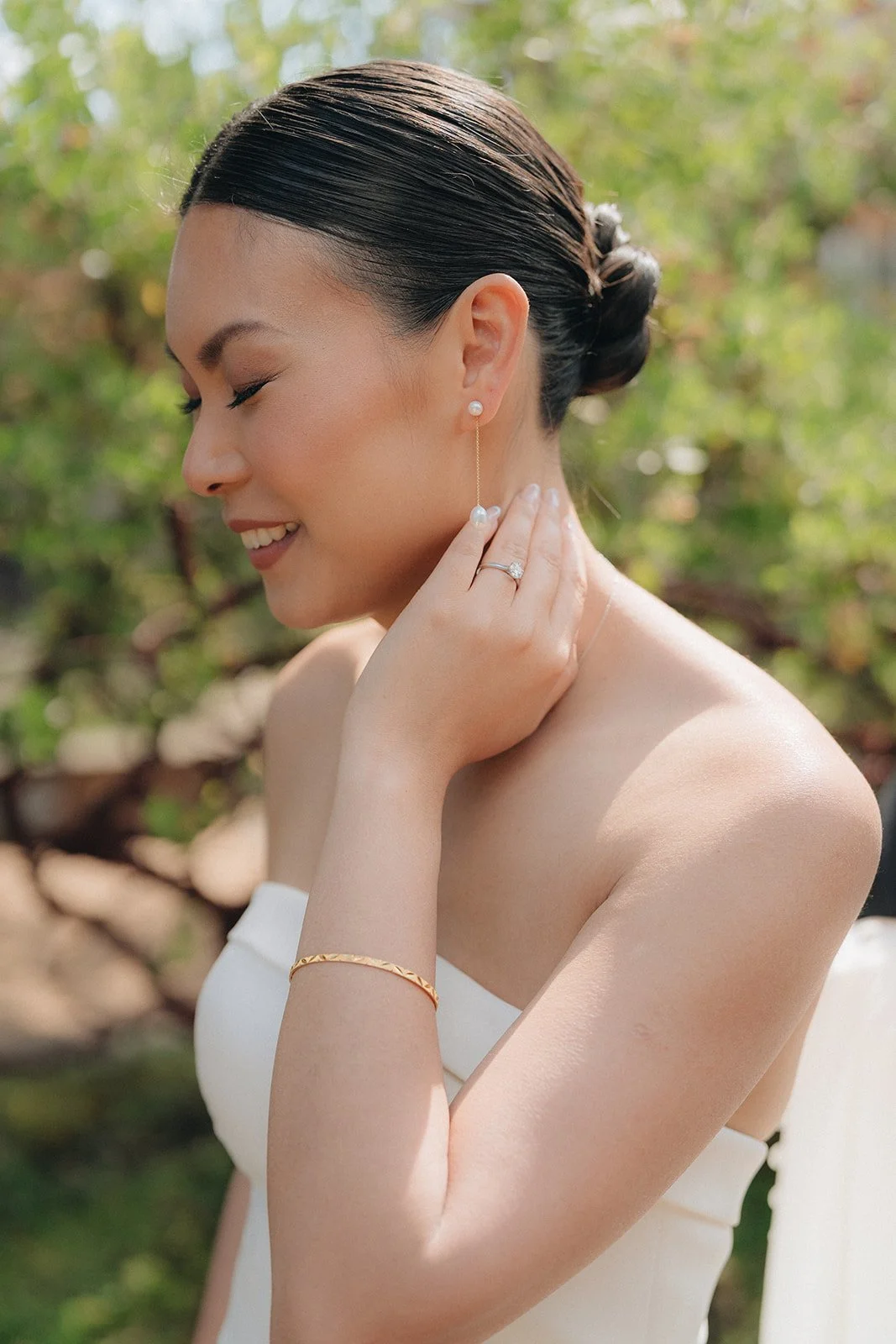 A woman with dark hair in a bun, wearing pearl earrings and a white strapless dress, smiling with her eyes closed outdoors.