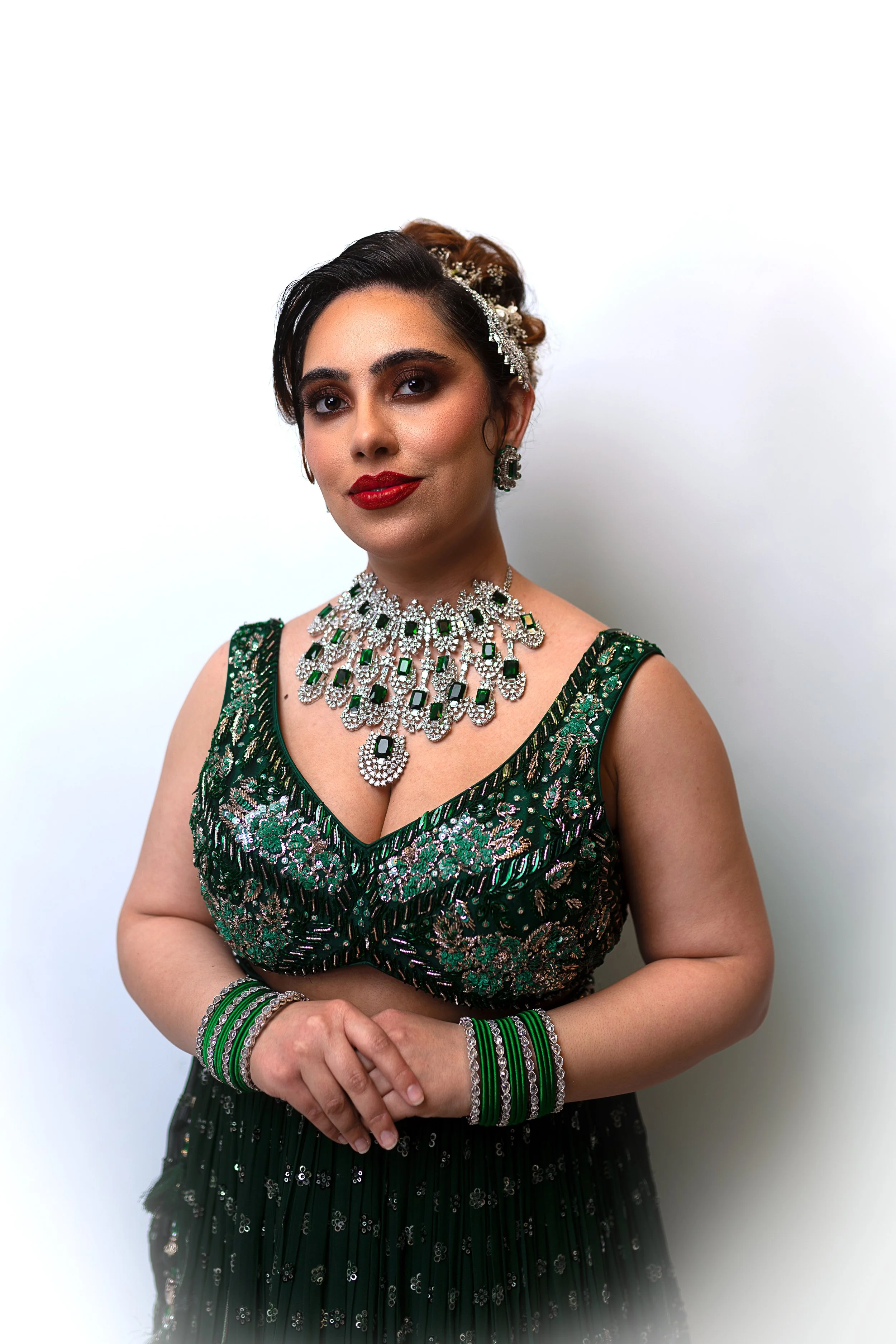 Woman dressed in a green and silver embroidered gown, wearing elaborate jewelry including a large pearl and emerald necklace, matching earrings, bracelets, and a headpiece with flowers, standing against a plain white background.