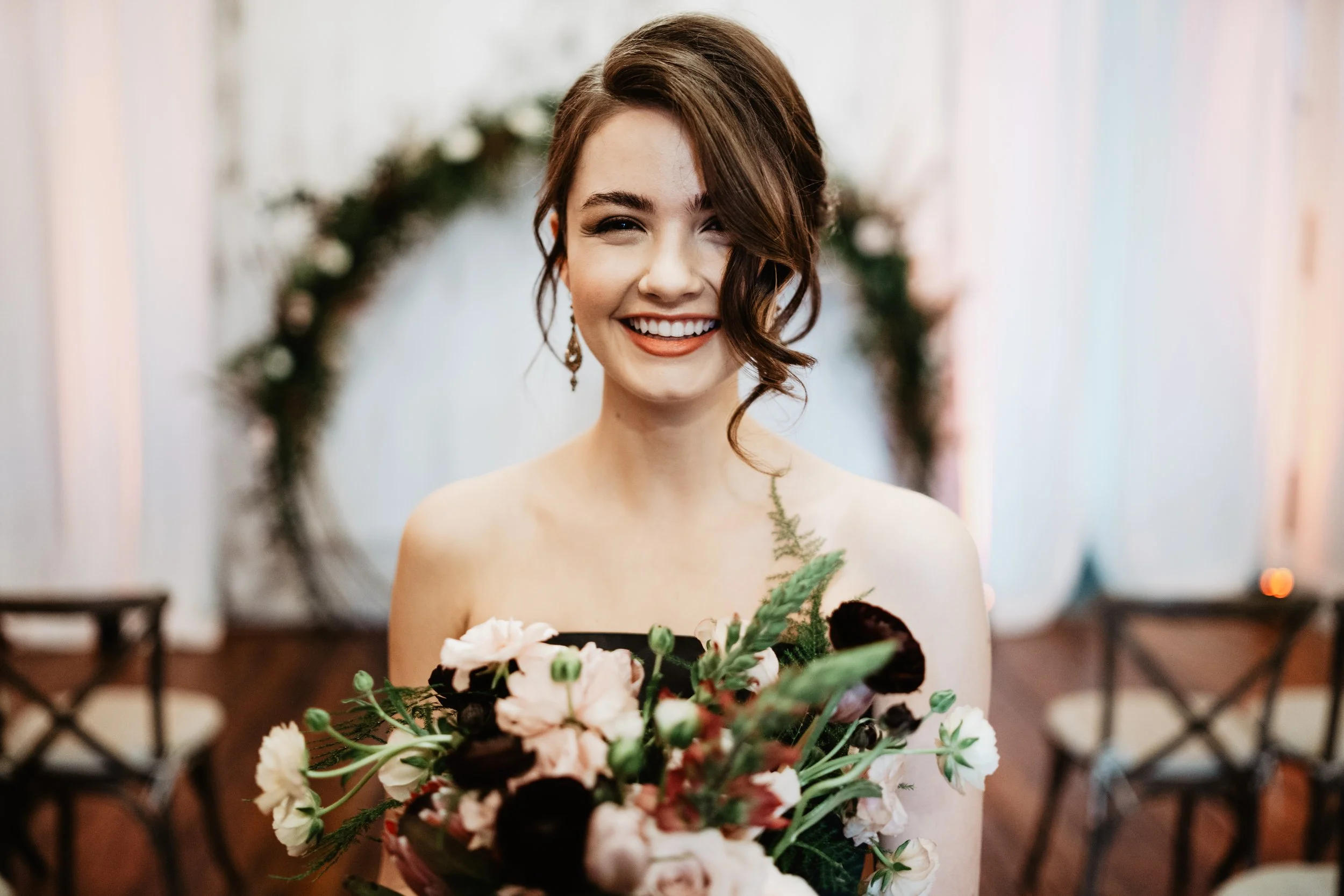 A woman in a wedding dress holding a bouquet of pink and white flowers, smiling with some of her hair hanging over her face, standing indoors in front of a blurred background with chairs and a green wreath.