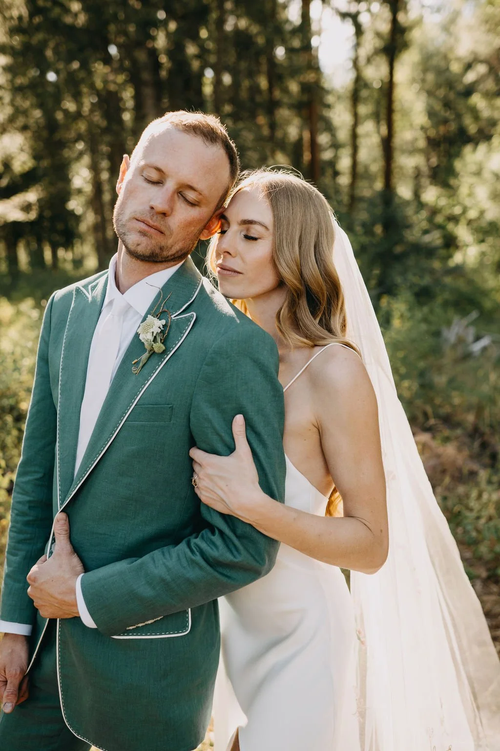 A bride and groom embrace outdoors during sunset, with the bride resting her head on the groom's shoulder, both with eyes closed, in a forest setting.