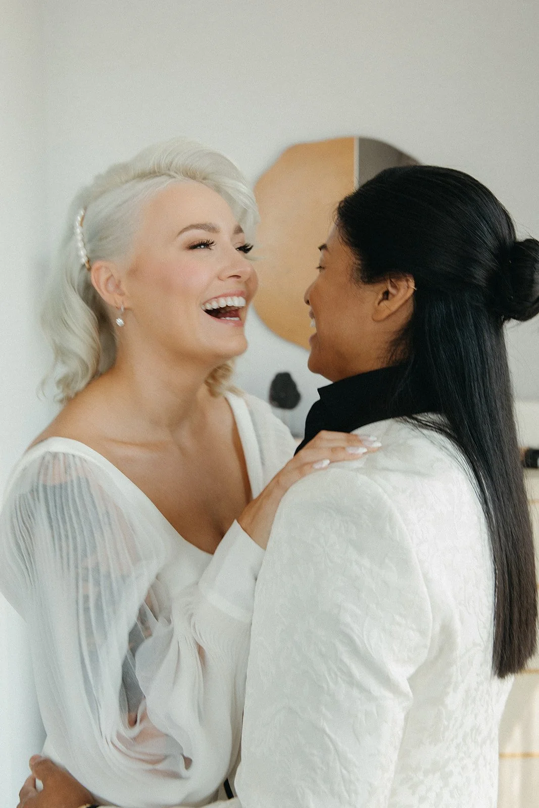 Two women laughing and smiling at each other in a close hug, one with platinum blonde hair and the other with black hair in an indoor setting.