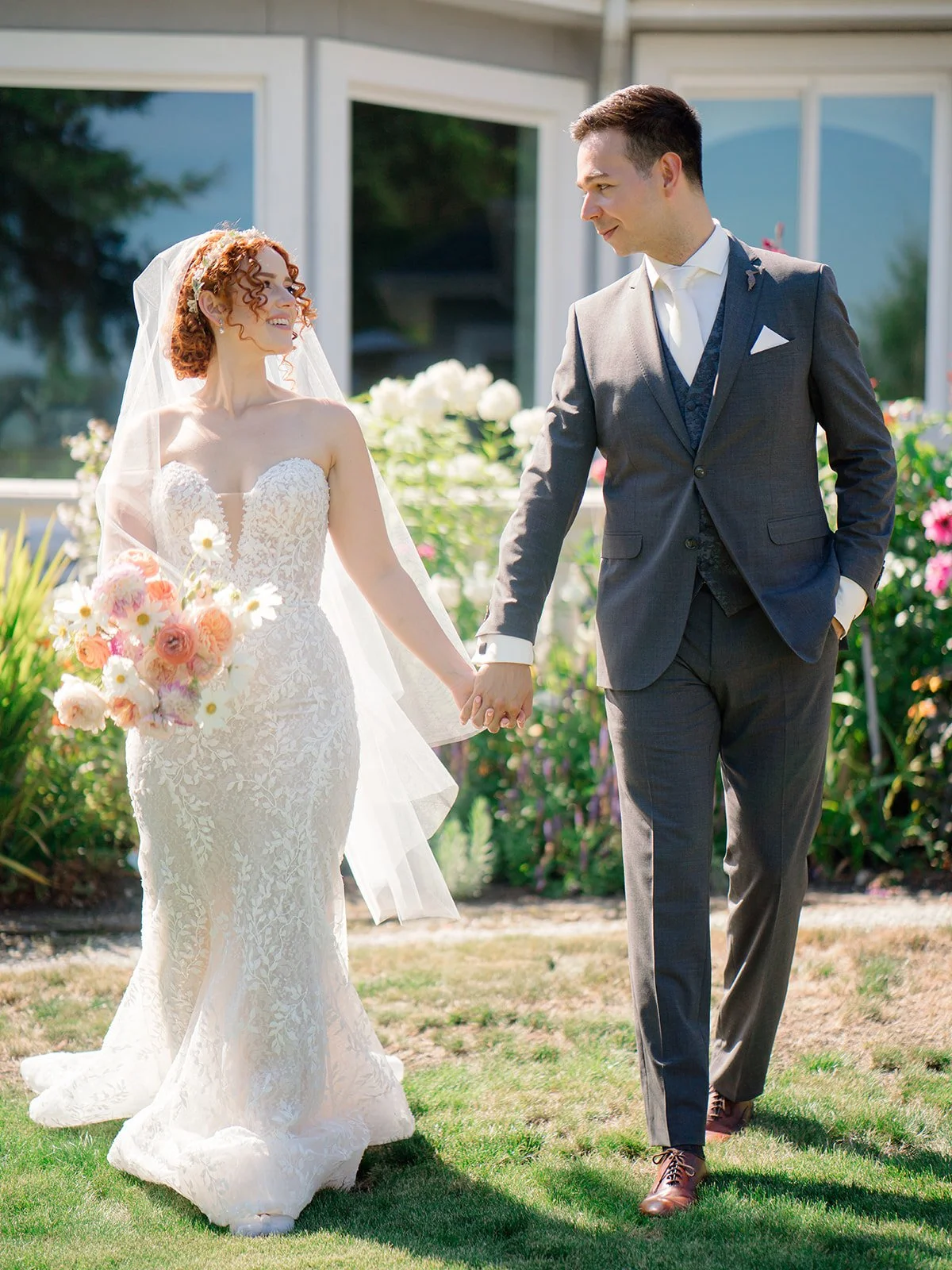 A bride and groom holding hands outdoors during their wedding, smiling at each other, with a garden and modern house in the background.
