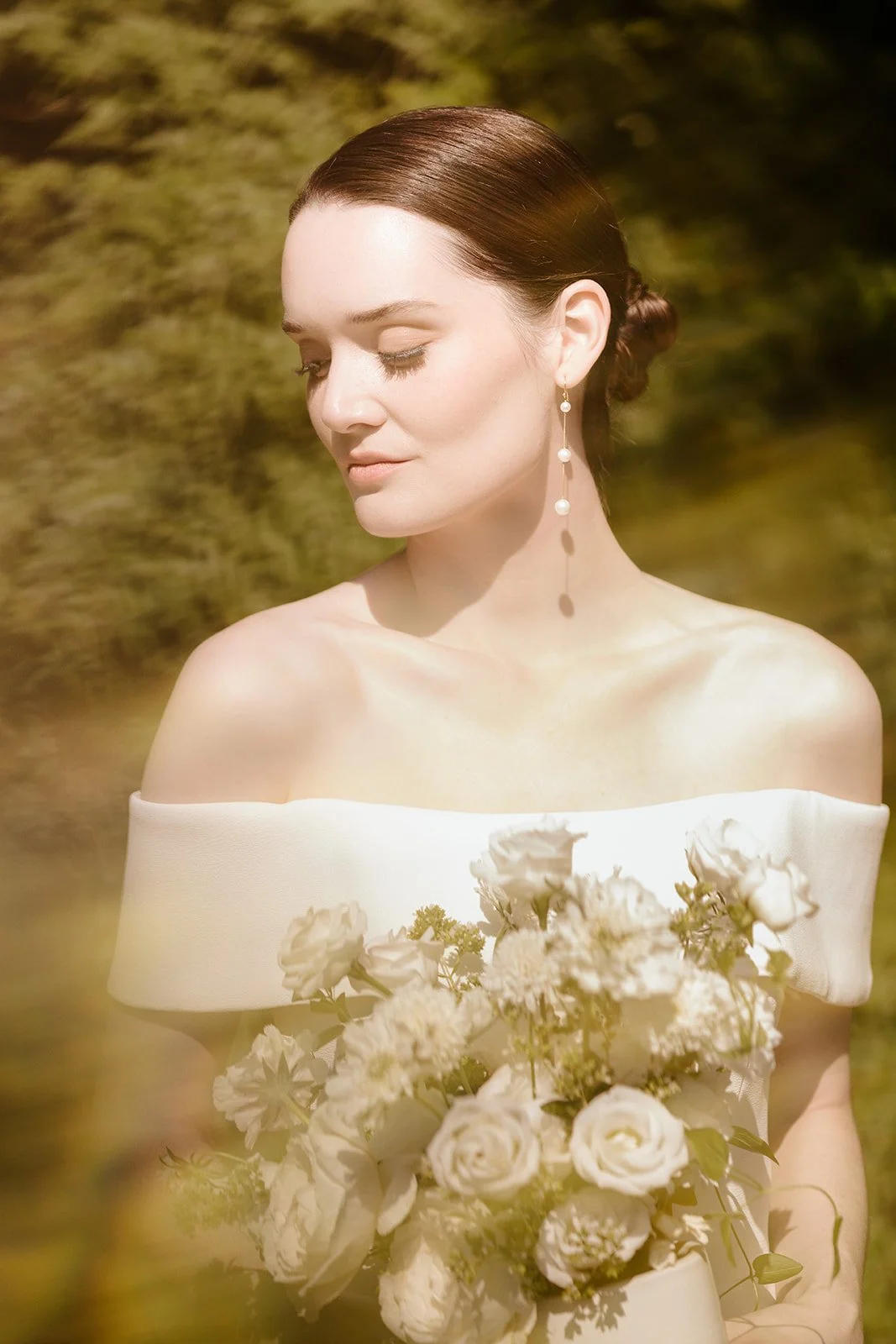 A woman with brown hair styled in an elegant updo, wearing pearl drop earrings and a white off-the-shoulder dress, holds a bouquet of white flowers outdoors with a blurred green background.