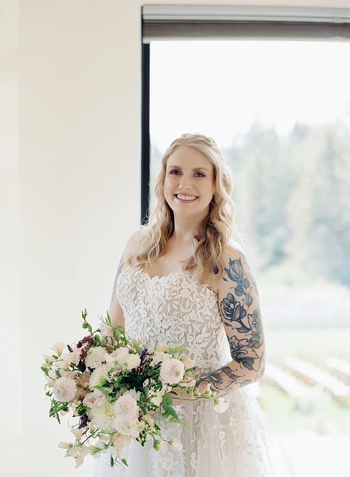 A smiling woman in a wedding dress holding a bouquet of flowers in front of a window.