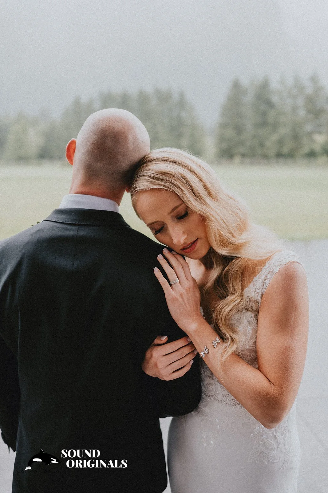 A bride and groom share an intimate moment outdoors, with the bride resting her head on the groom's shoulder, wearing a wedding gown and jewelry, against a blurred natural background.