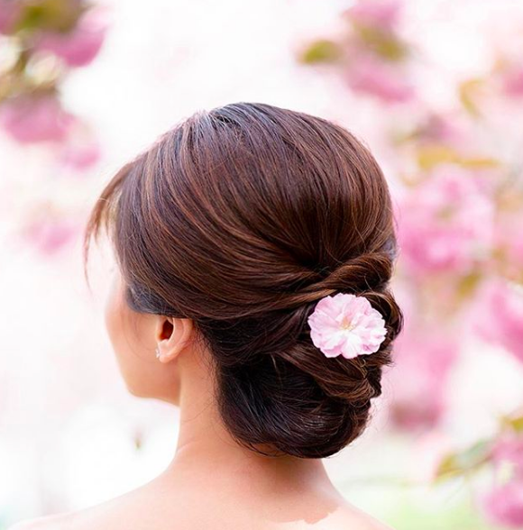 Back view of a woman's hairstyle with a pink flower accessories, outdoors with pink flowers in the background.