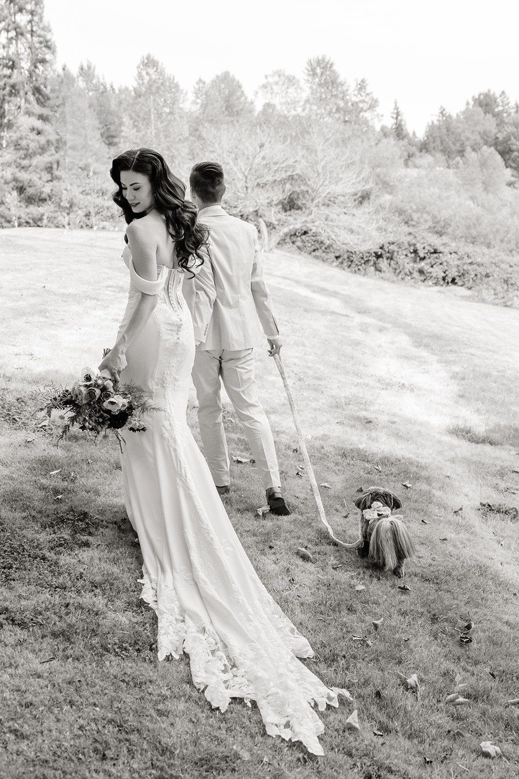 A black-and-white photo of a woman in a wedding dress holding a bouquet, and a man in a suit holding a small dog on a leash, outdoors on a grassy field with trees in the background.
Makeup and hair Stace Cho
Photographer Taylor Ochoa, Tminspired
Venu