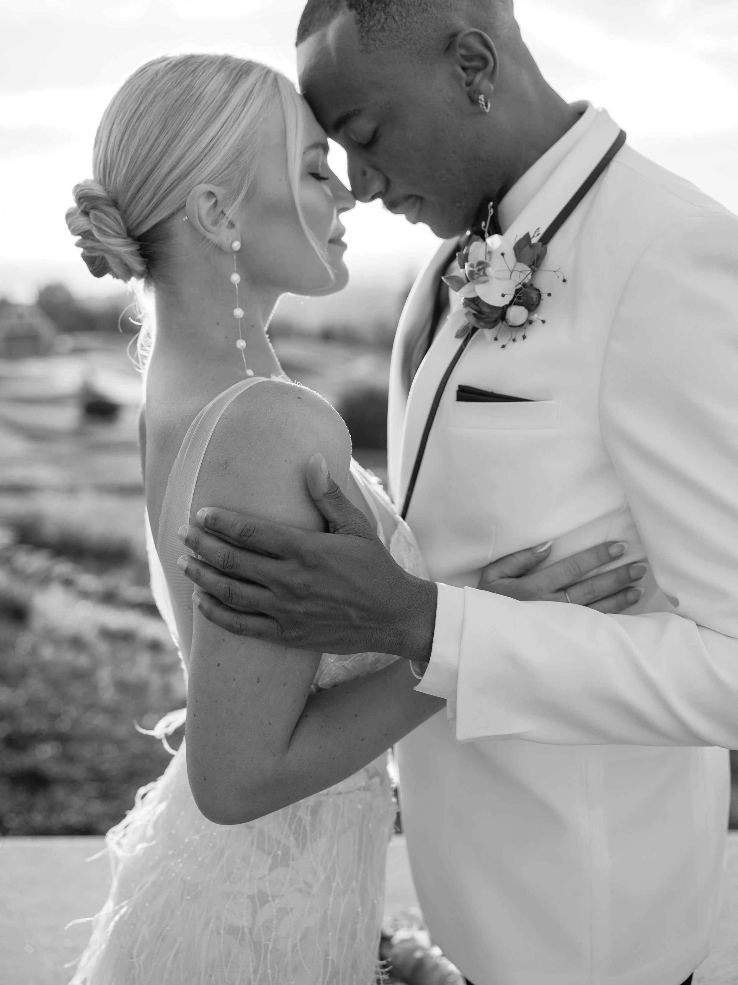 A black and white photo of a bride and groom standing very close with their foreheads touching, eyes closed, and smiling softly. The bride has light-colored hair styled in an updo, and the groom has dark skin and short hair with earrings. They are embracing each other, with the bride's hand on the groom's chest and the groom's arm around the bride's waist. The groom is wearing a white tuxedo with a boutonniere.