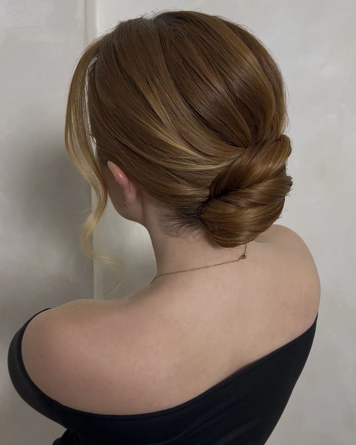 A woman with ginger hair styled in an elegant low bun with a wavy strand framing her face, wearing a black off-shoulder top and a delicate gold necklace, facing a plain light-colored wall.